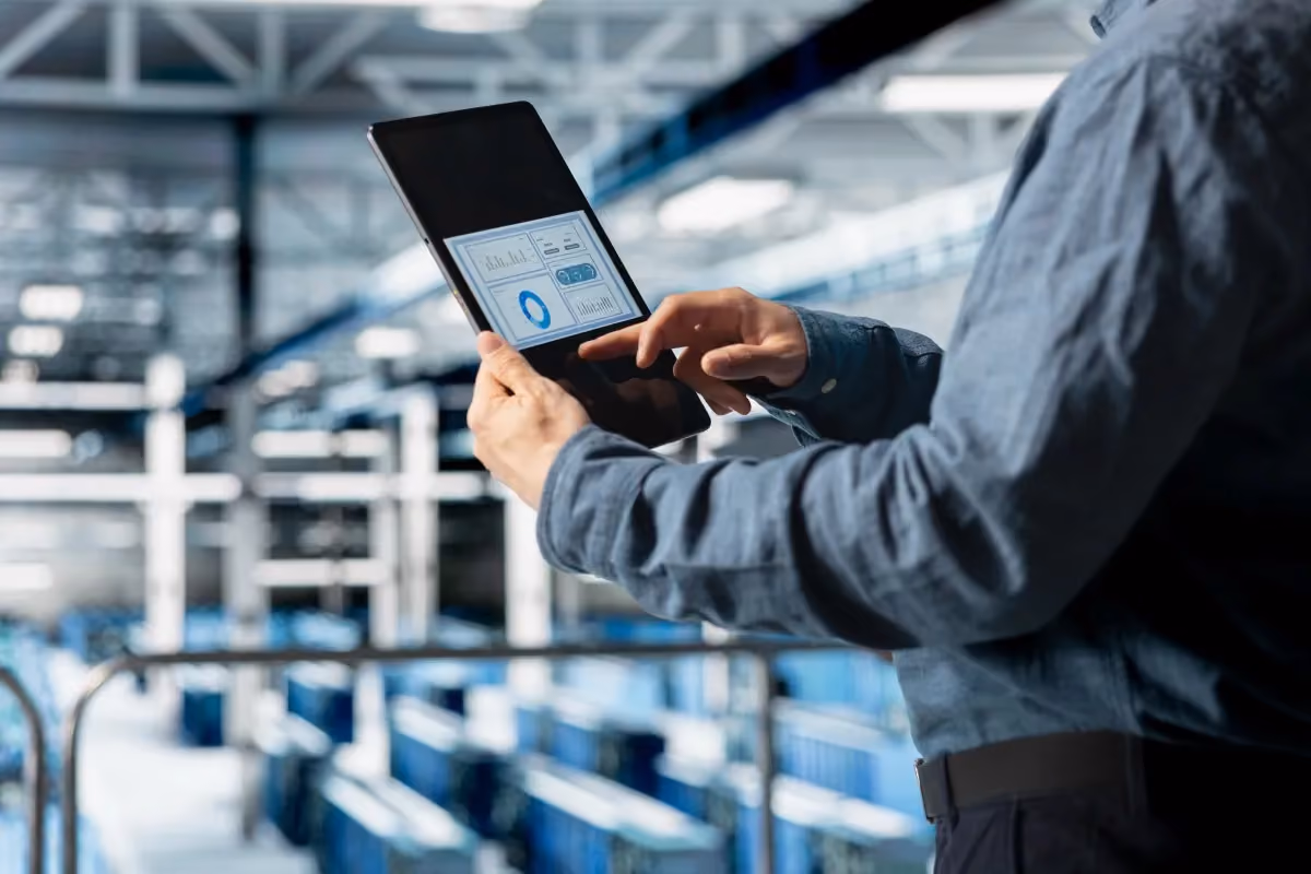 Person in blue shirt using a tablet displaying data charts in a modern industrial or warehouse setting.