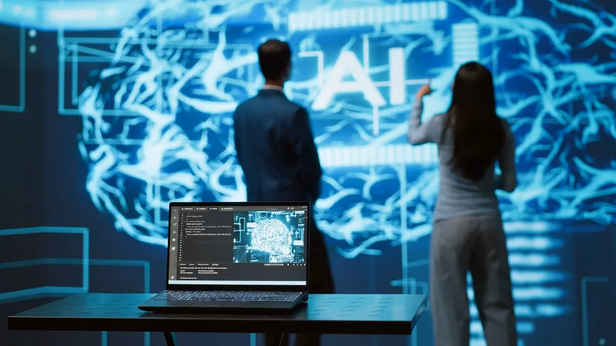 Laptop displaying code and a digital brain illustration on a table with two professionals interacting with a large AI neural network display in the background.