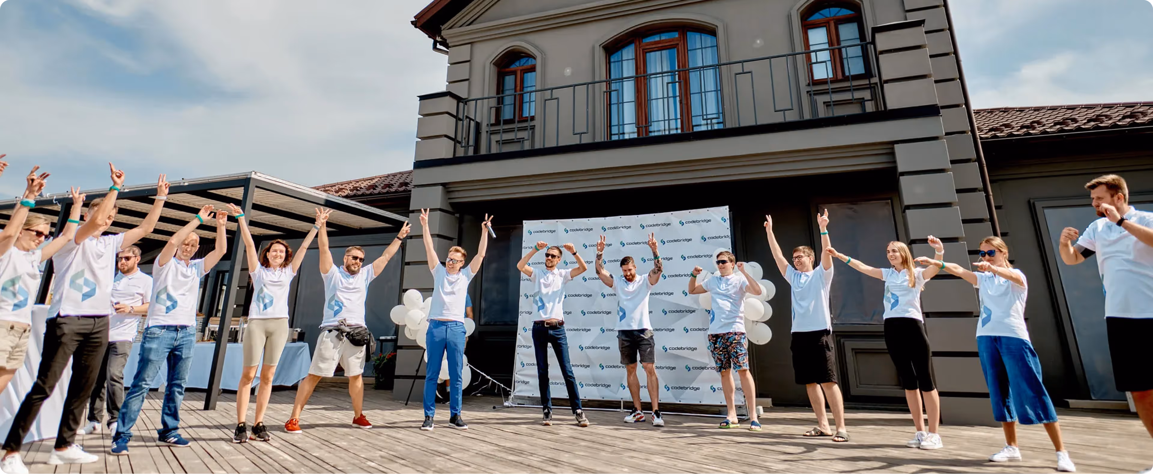 Group of people wearing white shirts celebrating outdoors with arms raised on a wooden deck in front of a building with a Codebridge banner.