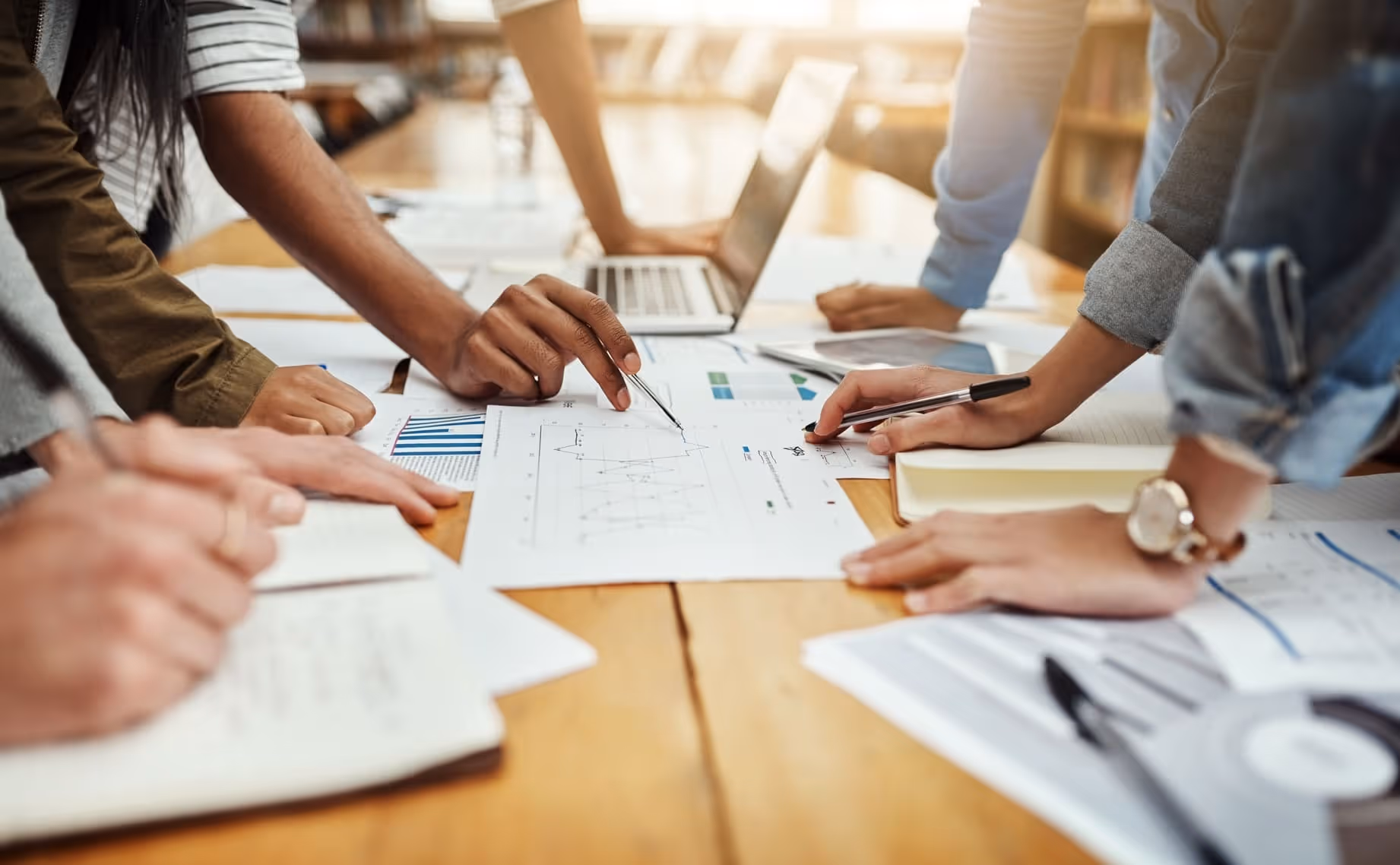 Close-up of people collaborating over documents with charts and graphs on a wooden table, with a laptop in the background.
