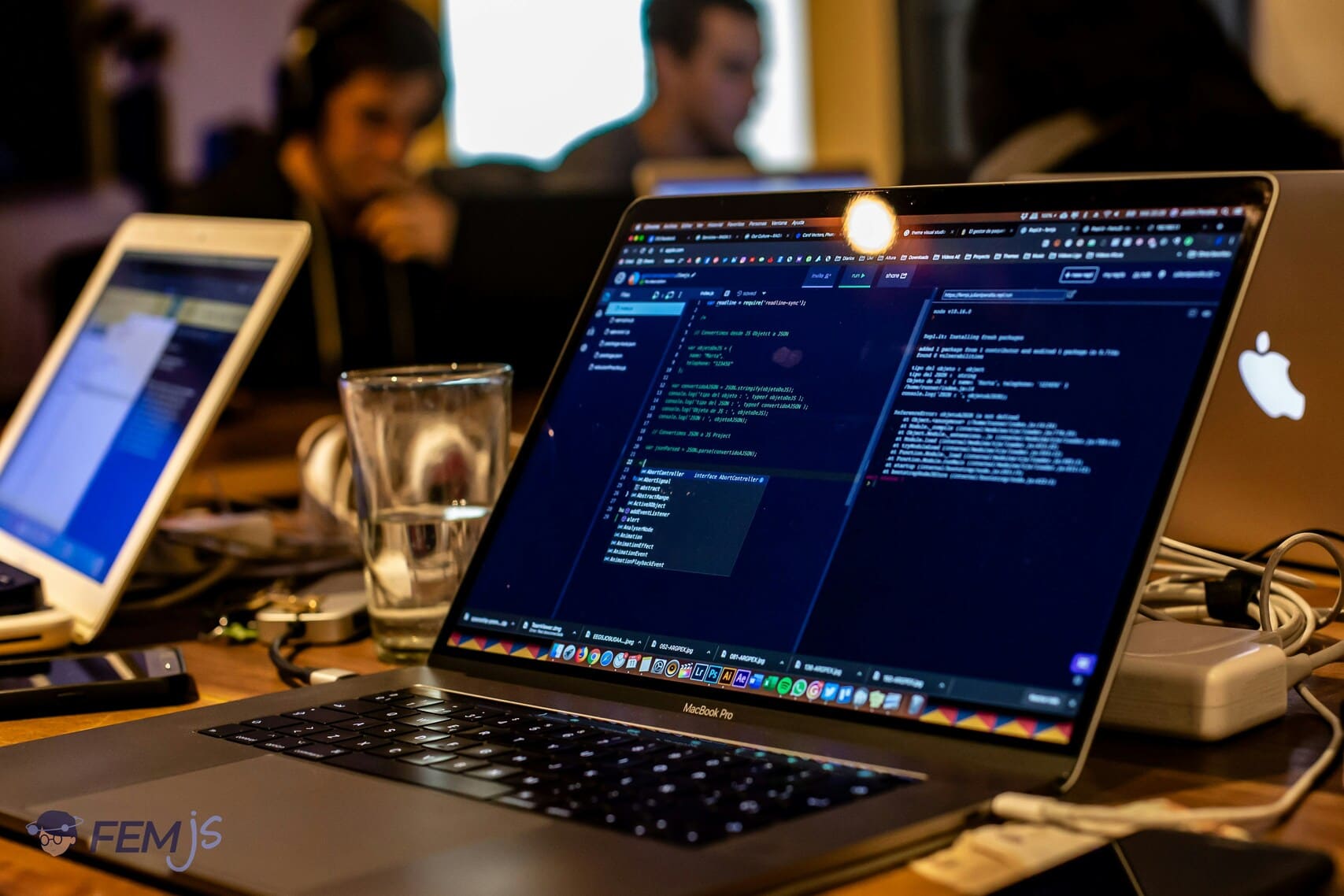 MacBook Pro laptop on a desk displaying coding software with two blurred people working in the background.
