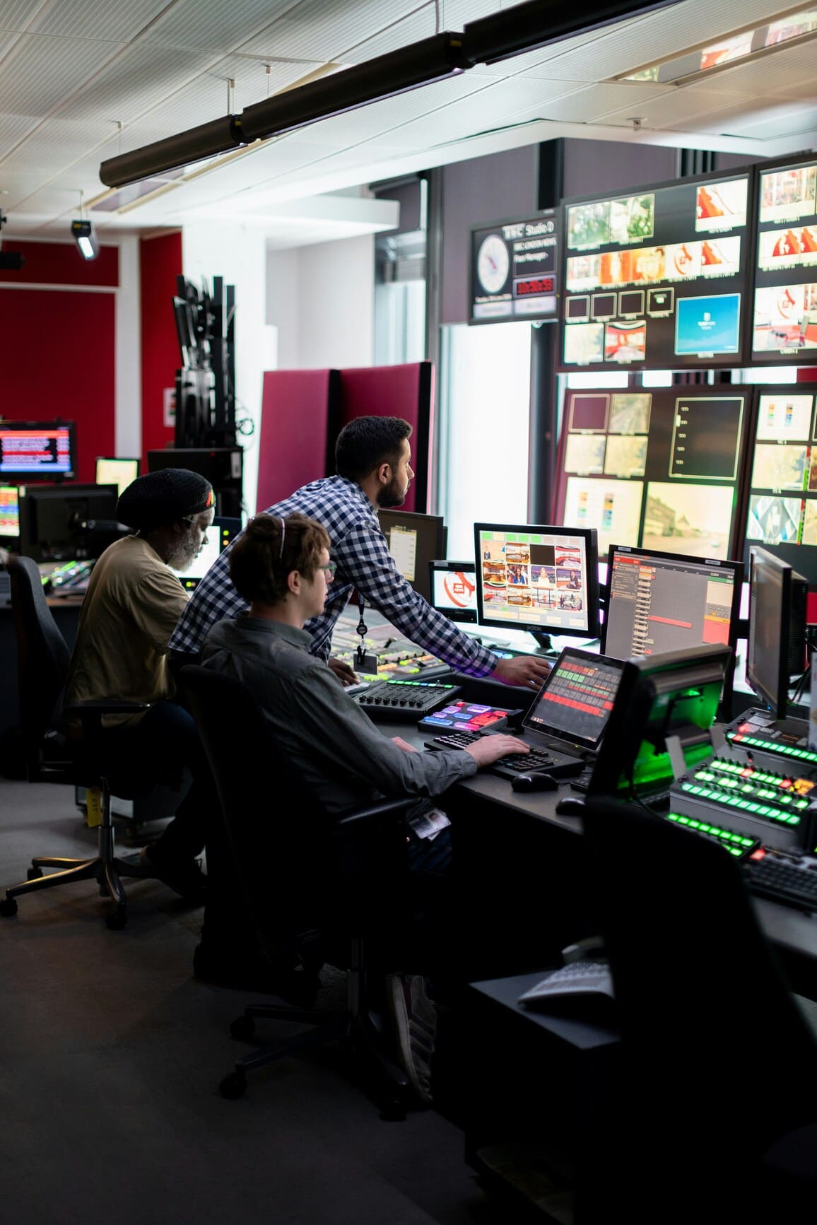 Three men operating multiple monitors and control panels in a broadcast control room.