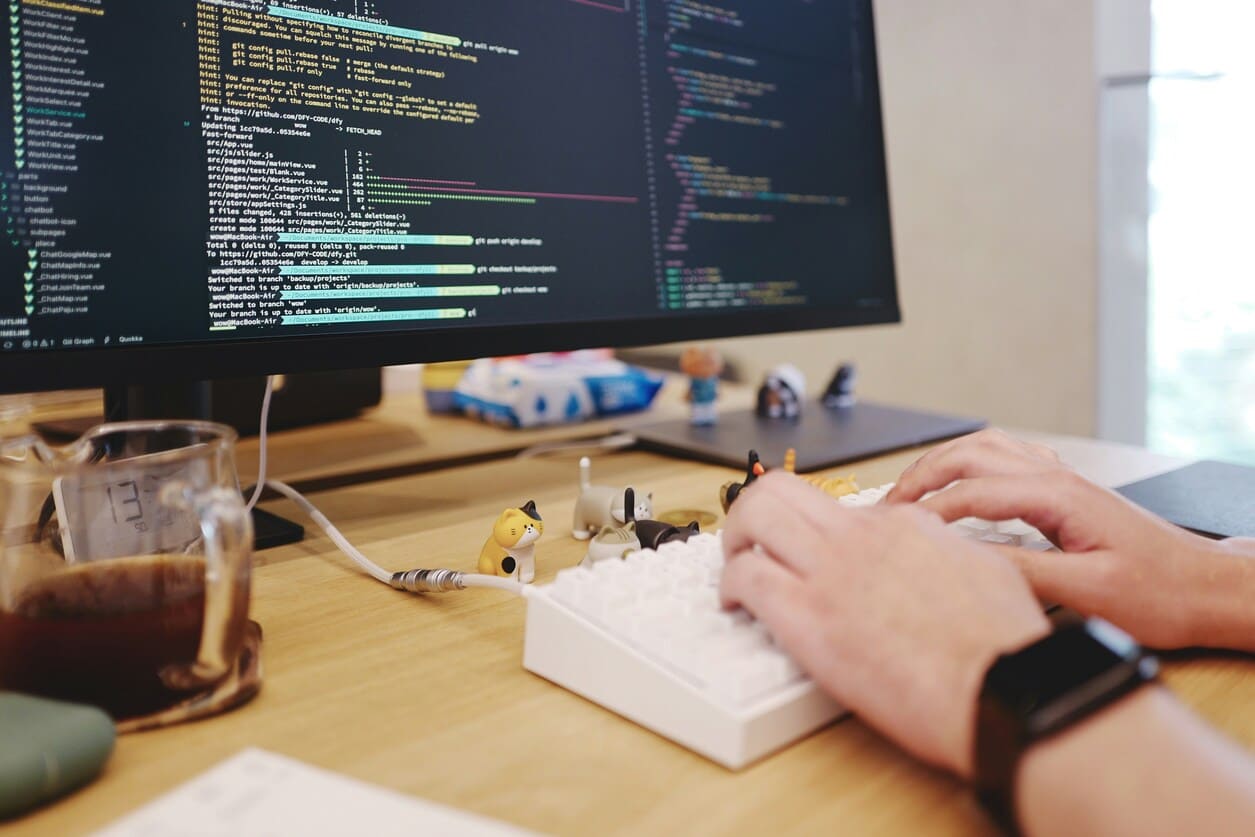 Person typing on a white keyboard decorated with small cat figurines, with a computer screen displaying code and a glass of coffee on the desk.