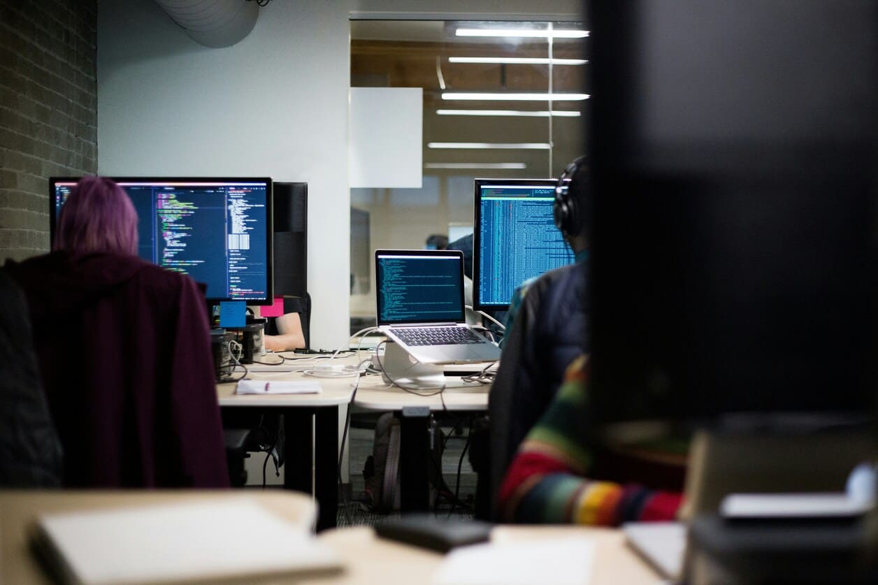 People working at desks with computer screens displaying code and data in an office setting.