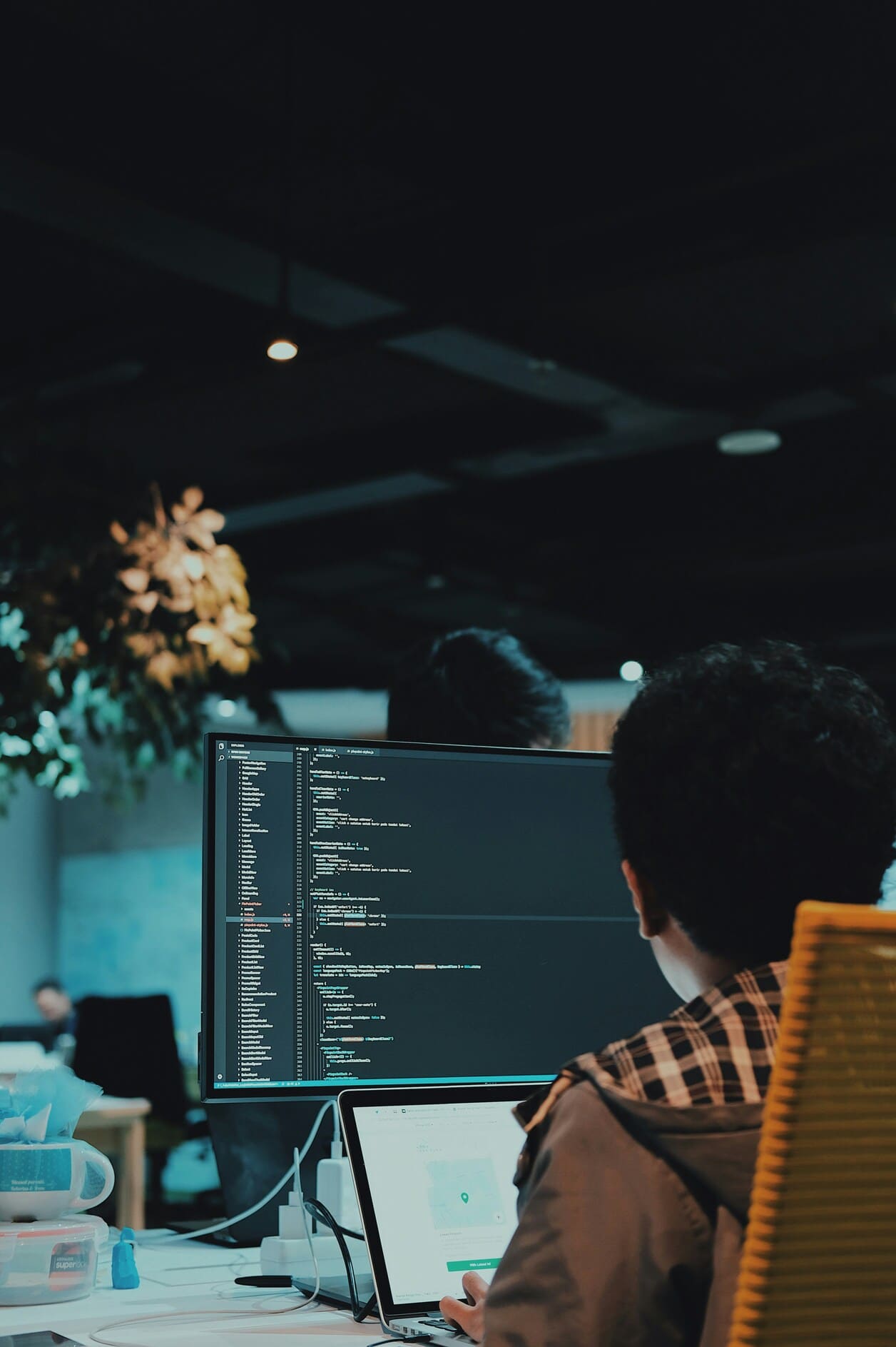 Person sitting at a desk working on a laptop with a large monitor displaying programming code in a dimly lit office.