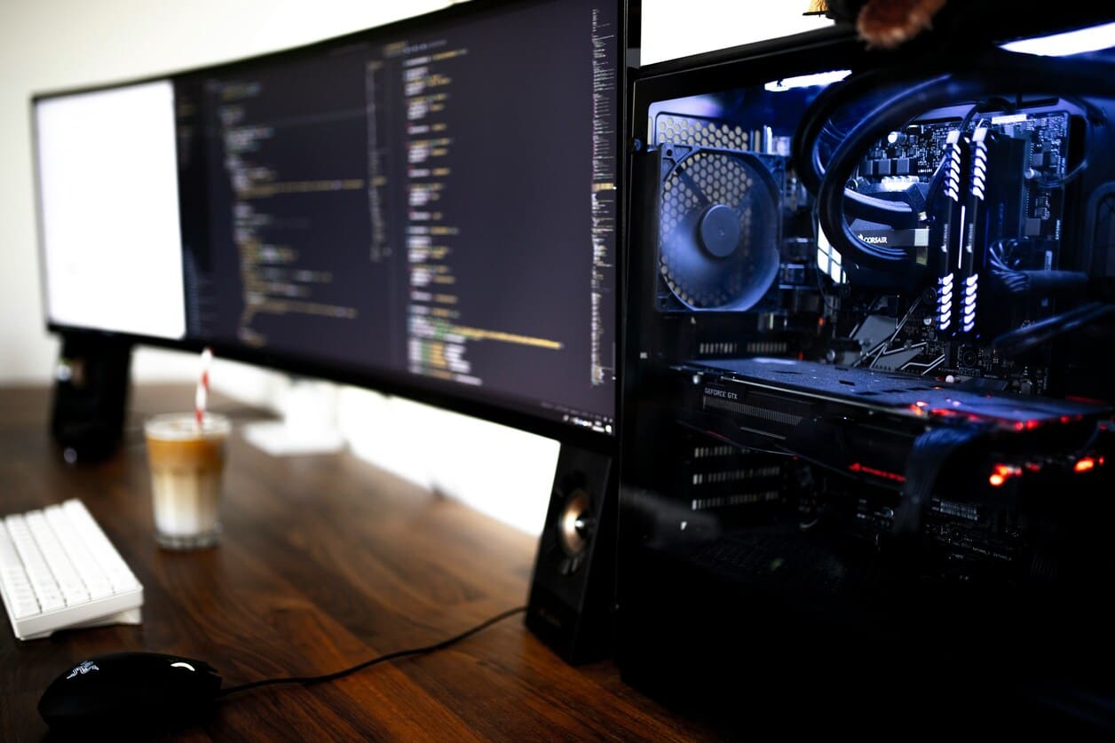 Modern desktop computer setup with a curved monitor displaying code, a mechanical keyboard, mouse, and iced coffee on a wooden desk.