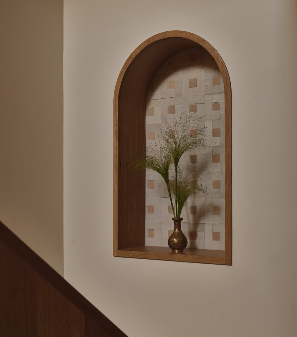 Wall niche with wooden frame holding a brass vase containing delicate green grass stems against a tiled background with square patterns.