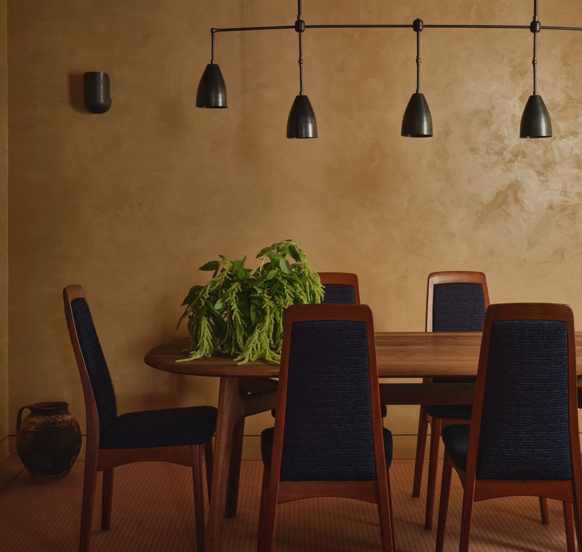 Dining area with wooden table surrounded by five dark-cushioned chairs, a green plant centerpiece, and four black pendant lights against a beige textured wall.