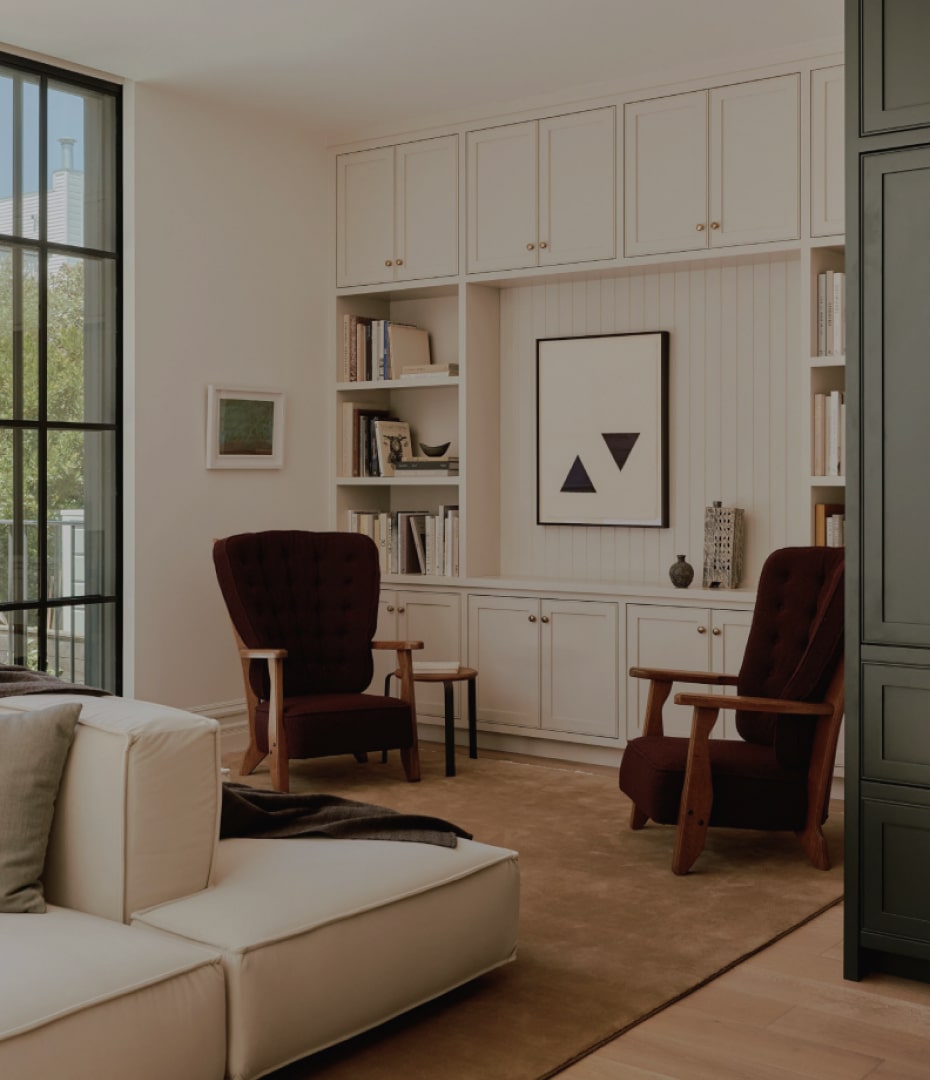 Cozy living room with two dark cushioned wooden armchairs, white built-in shelves with books and decor, a beige sectional sofa, and large window showing greenery outside.