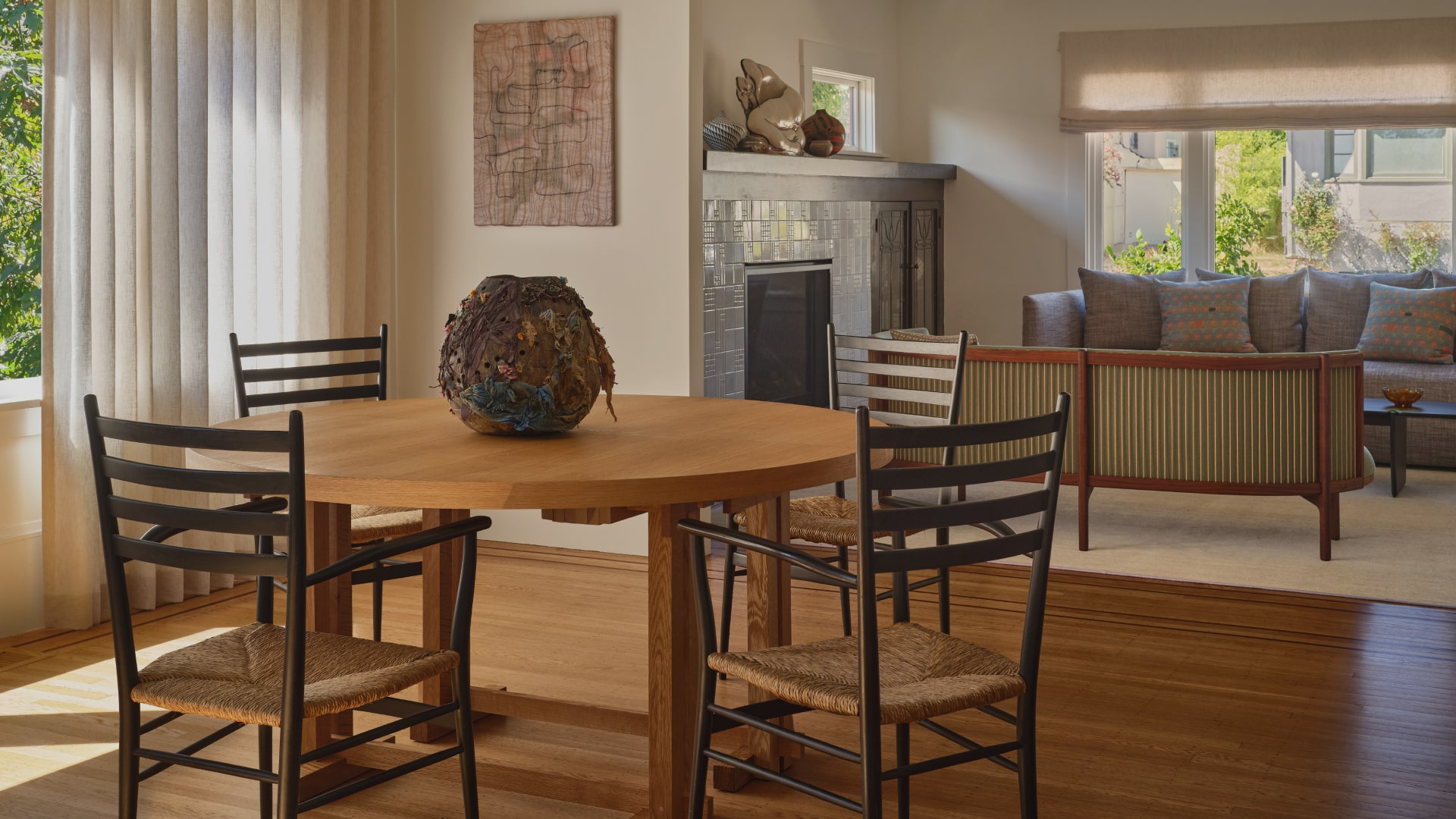 Sunlit dining area with a round wooden table surrounded by four black ladder-back chairs with woven seats, a decorative vase on the table, and a living room with sofa and fireplace in the background.