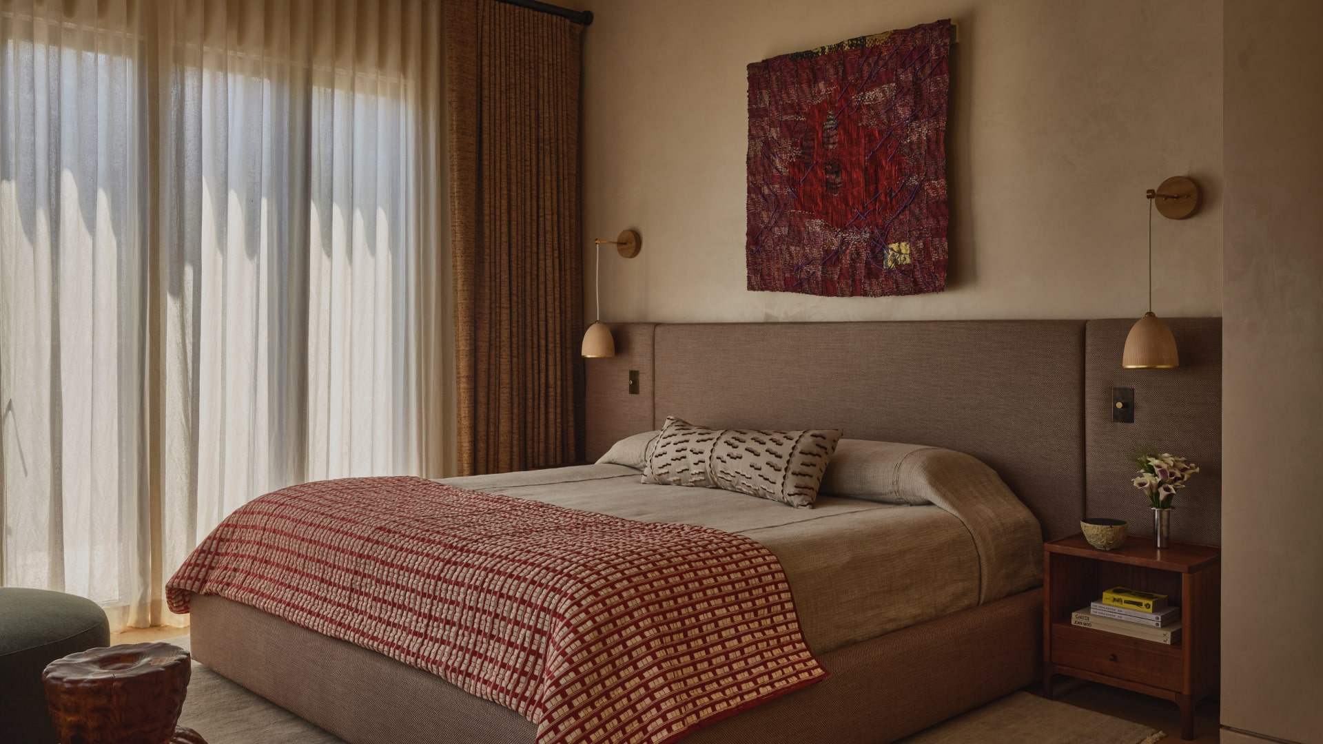 Modern bedroom with beige bedspread, red patterned blanket, matching pillows, wooden nightstand with flowers, and sheer curtains.
