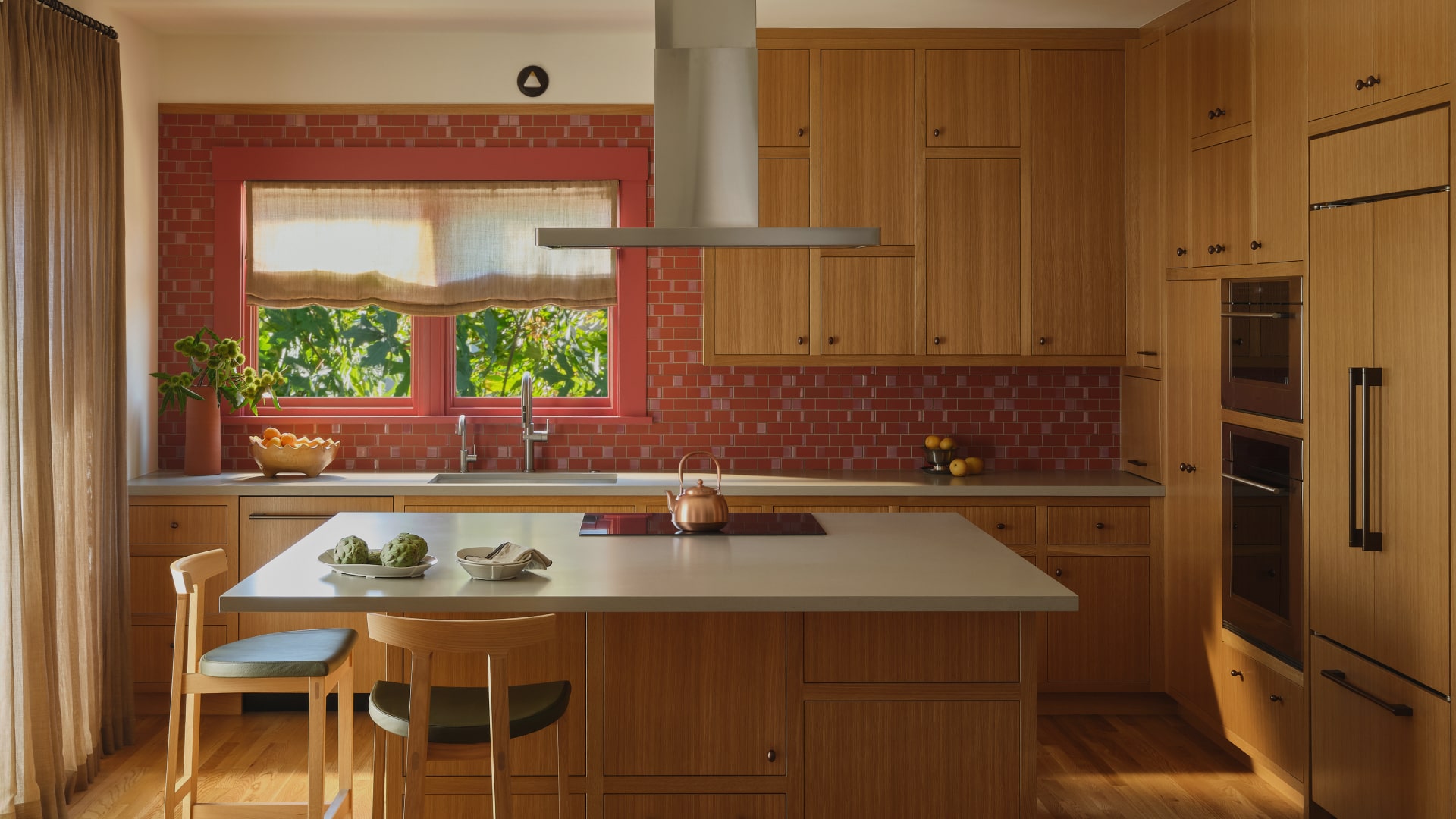 Modern kitchen with wooden cabinets, red tile backsplash, island with cooktop and copper kettle, and a window with beige curtains.