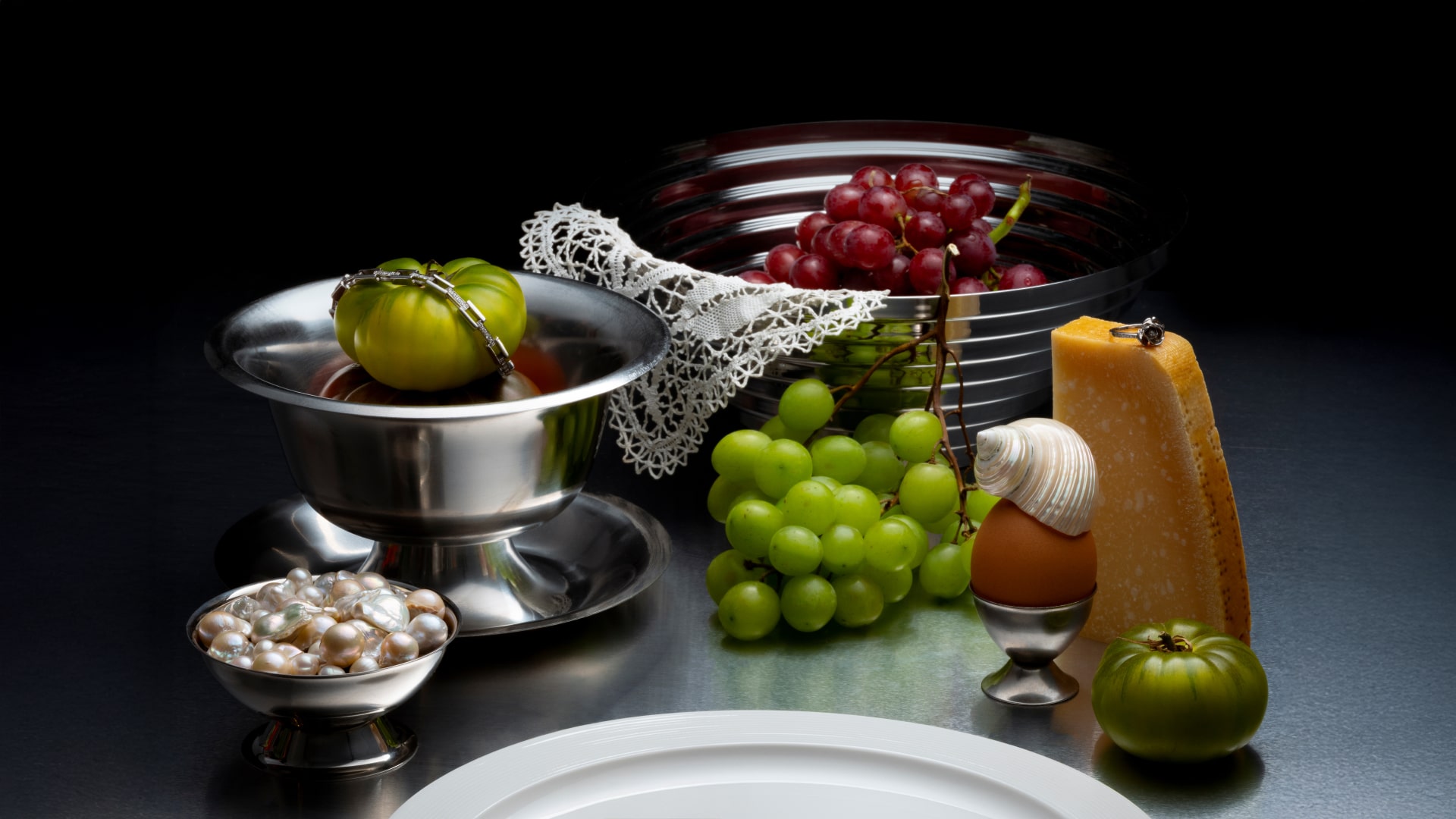 Still life with green and red grapes, green tomatoes, wedge of cheese with a ring, pearls in a silver bowl, egg with a seashell on top, and lace on metallic dishes.
