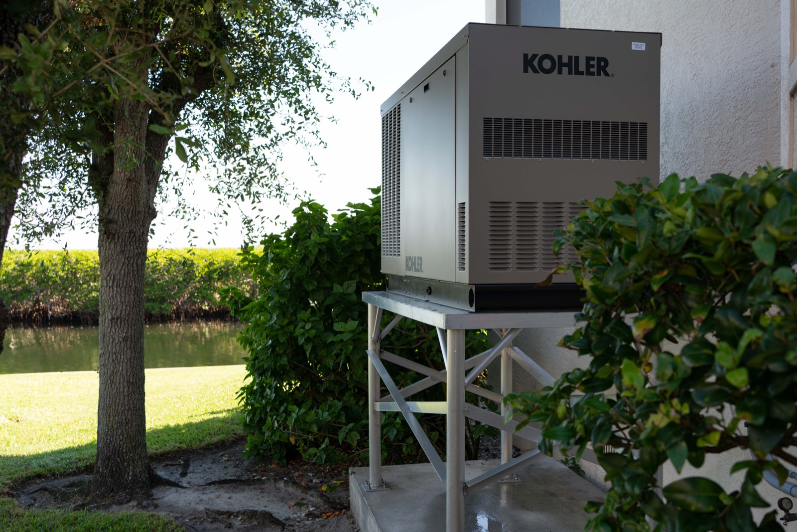A Kohler standby generator installed on a welded aluminum elevation stand bolted to a concrete pad, with a canal running behind the property.