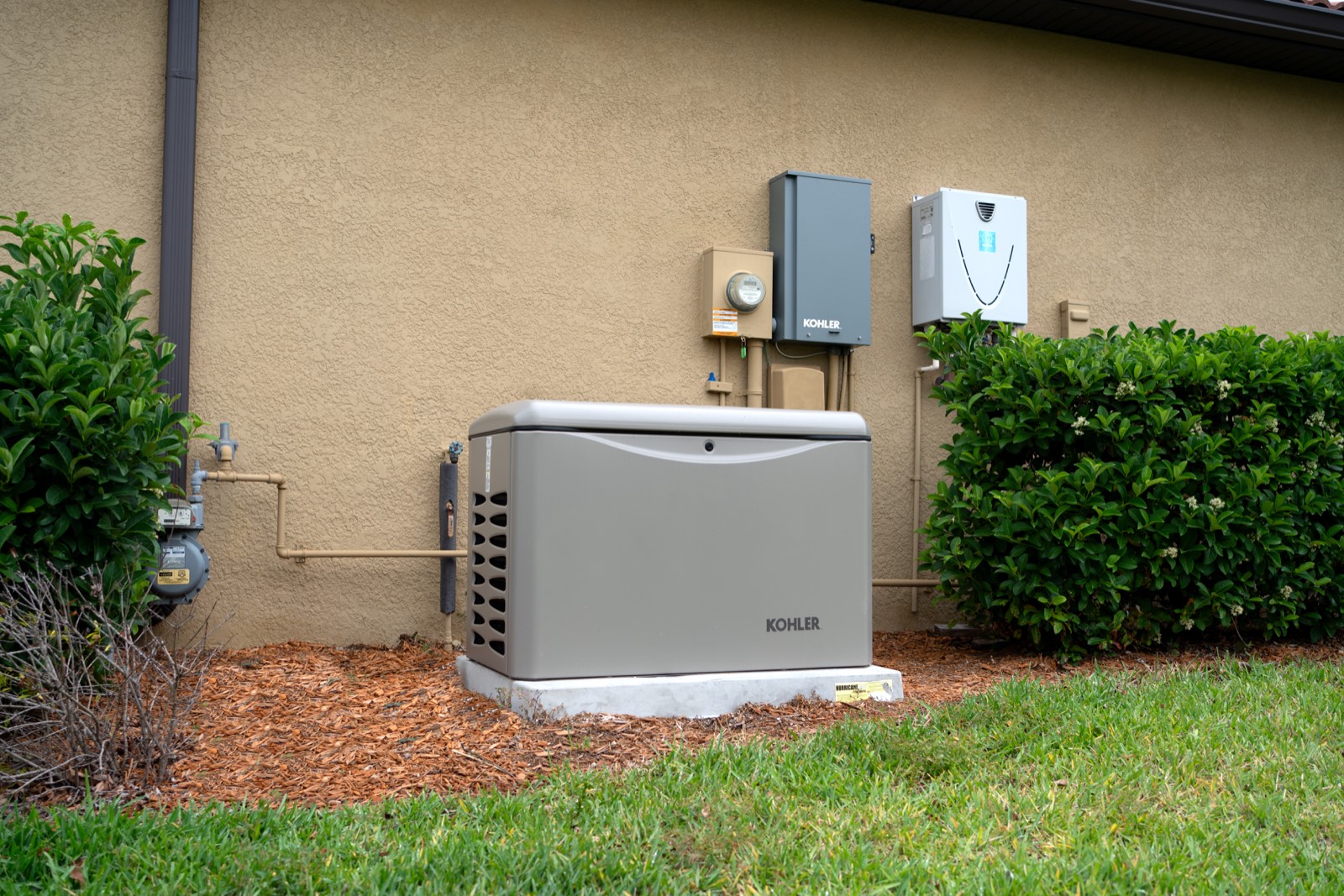 A completed Kohler generator installation on a concrete pad, with the gas meter and yellow gas line at left and the electric meter, Kohler transfer switch and tankless water heater mounted on the wall behind.
