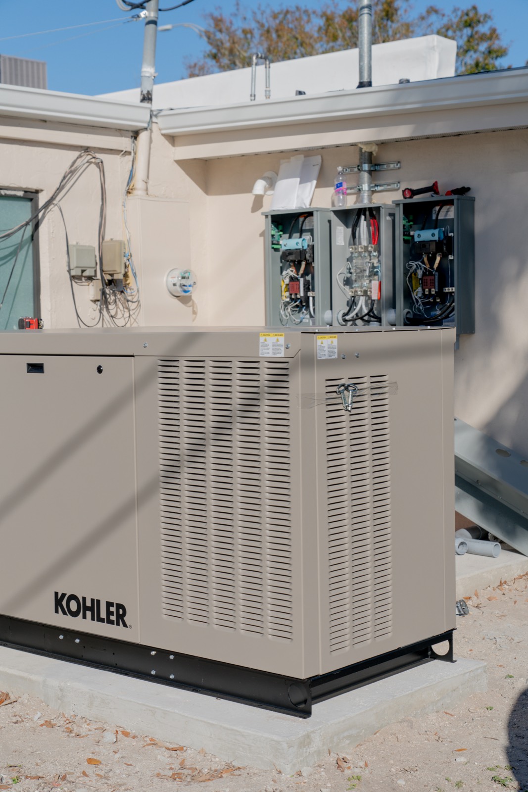 A newly installed Kohler standby generator on a concrete pad, with three transfer-switch enclosures open above it showing breakers and conductors mid-installation.