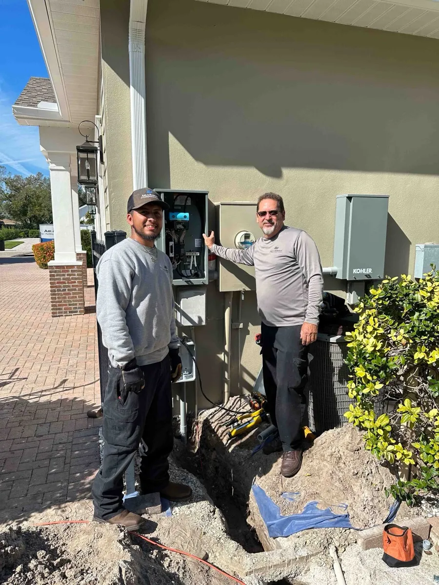 Two Air & Energy technicians beside an open transfer switch and electric meter on the side of a house, with a freshly dug trench for the generator's lines at their feet
