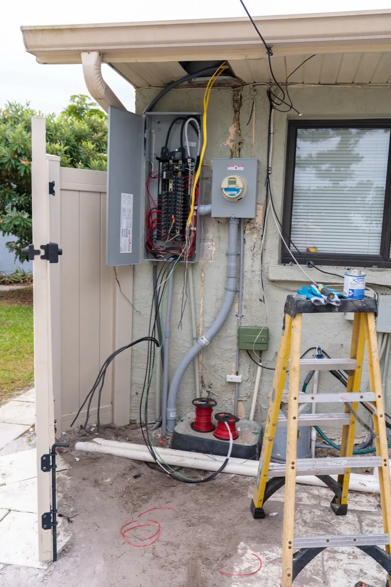 An open outdoor electrical panel and electric meter on the side of a house during a repair, with new wire spools and a ladder in front