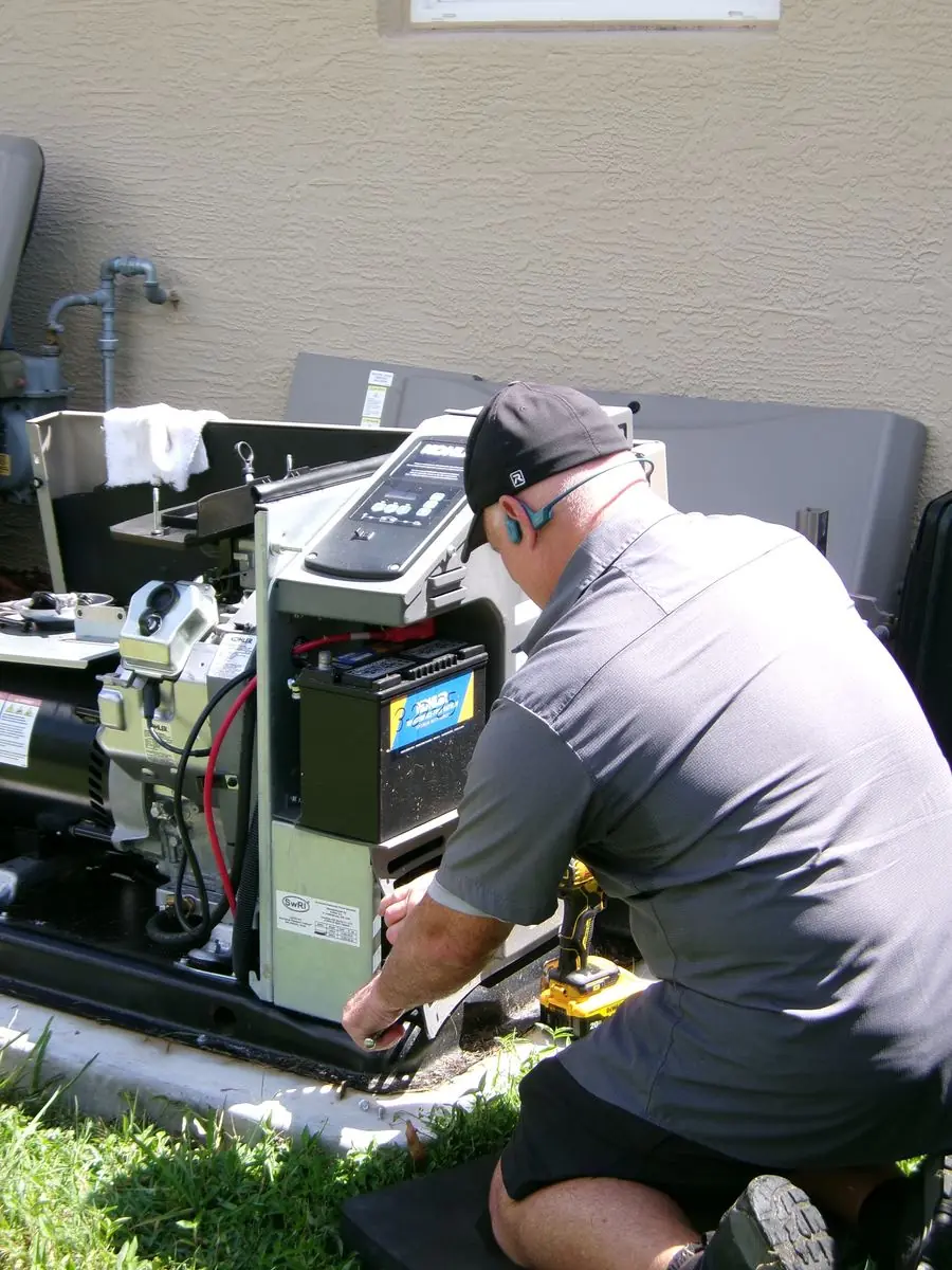 An Air & Energy technician kneels beside an air-cooled Kohler generator with its enclosure removed, working below the controller and battery