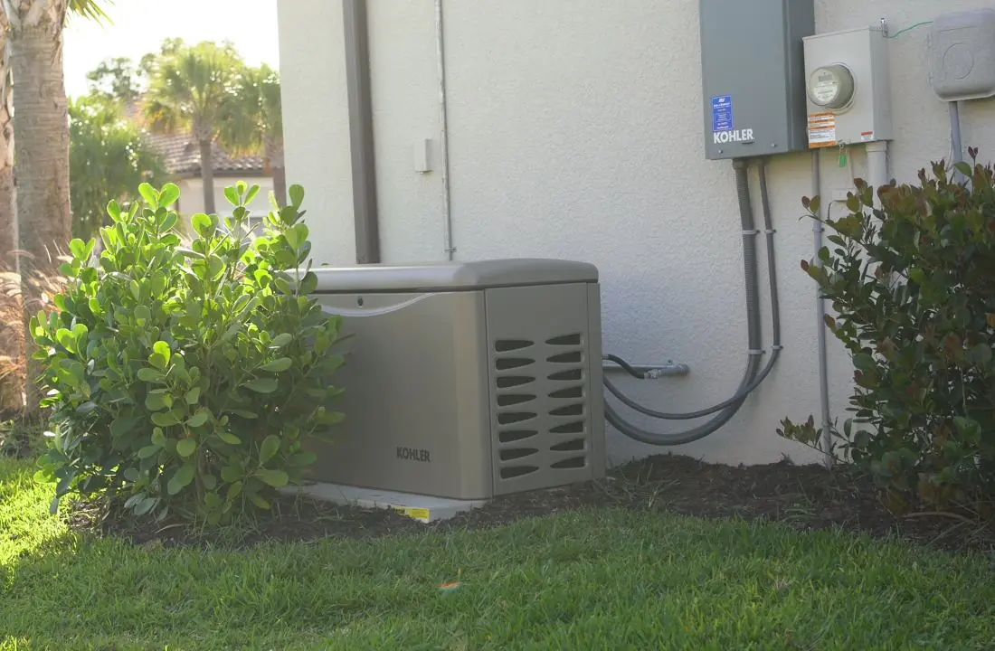 A Kohler home standby generator on its pad in a planted side yard, with the Kohler transfer switch and electric meter on the wall above