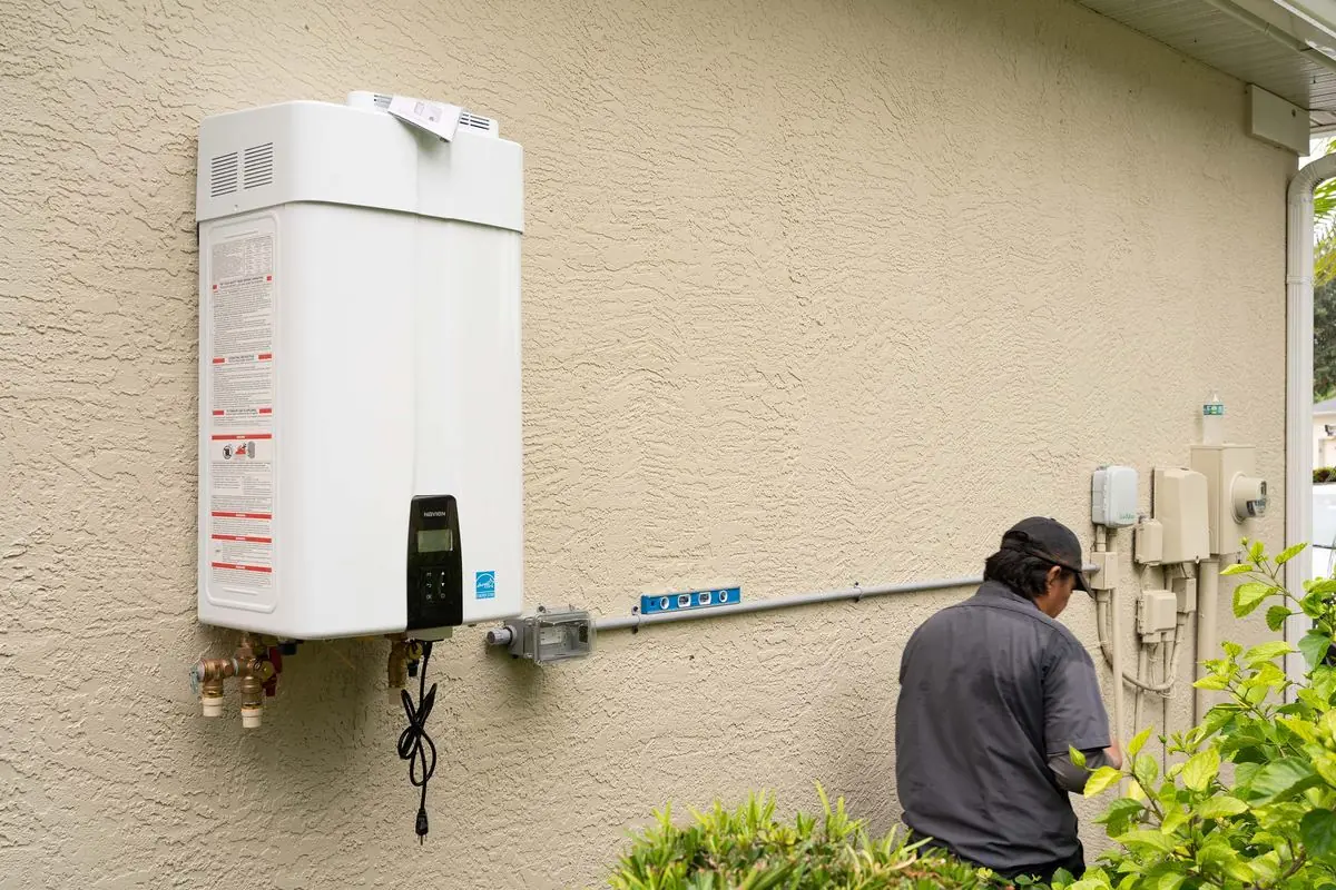 Air & Energy technician working at an outdoor electrical box beside a newly installed tankless water heater on a stucco wall