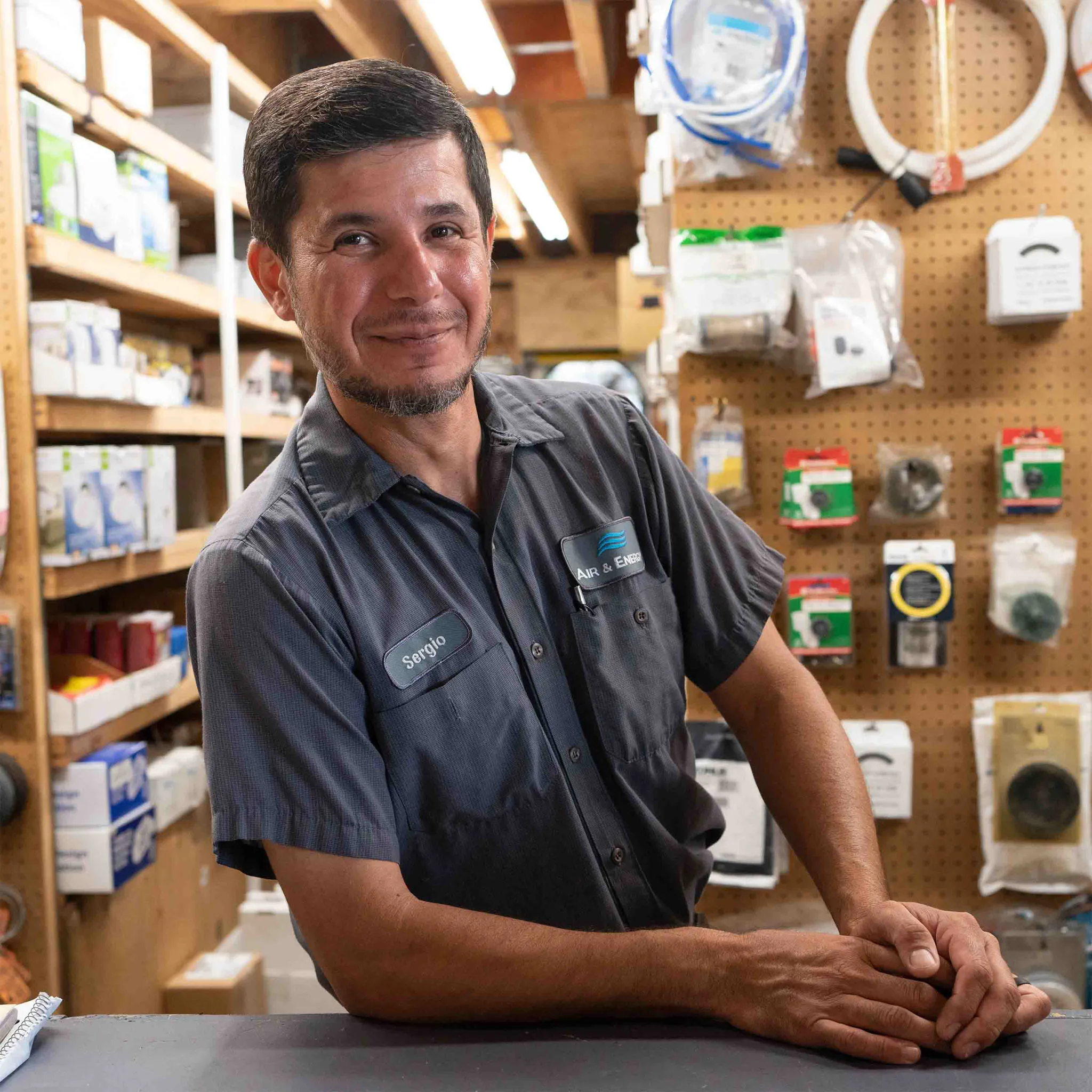 An Air & Energy team member in a company uniform smiling at the parts counter, with shelves of parts behind him