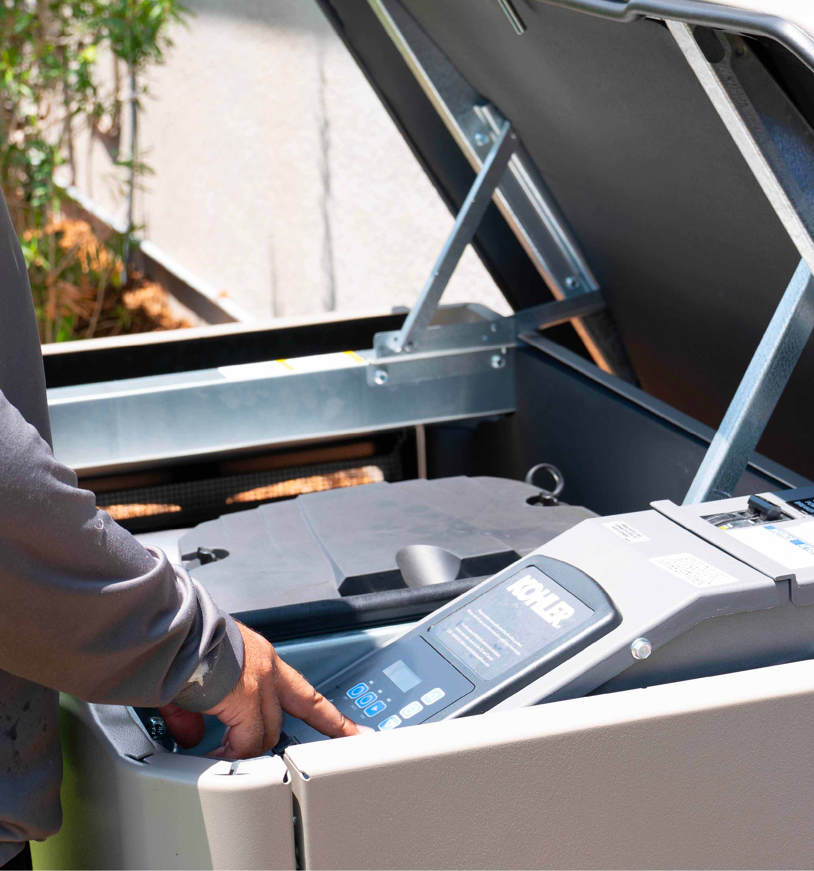 Person operating a Kohler generator control panel inside an open generator enclosure.