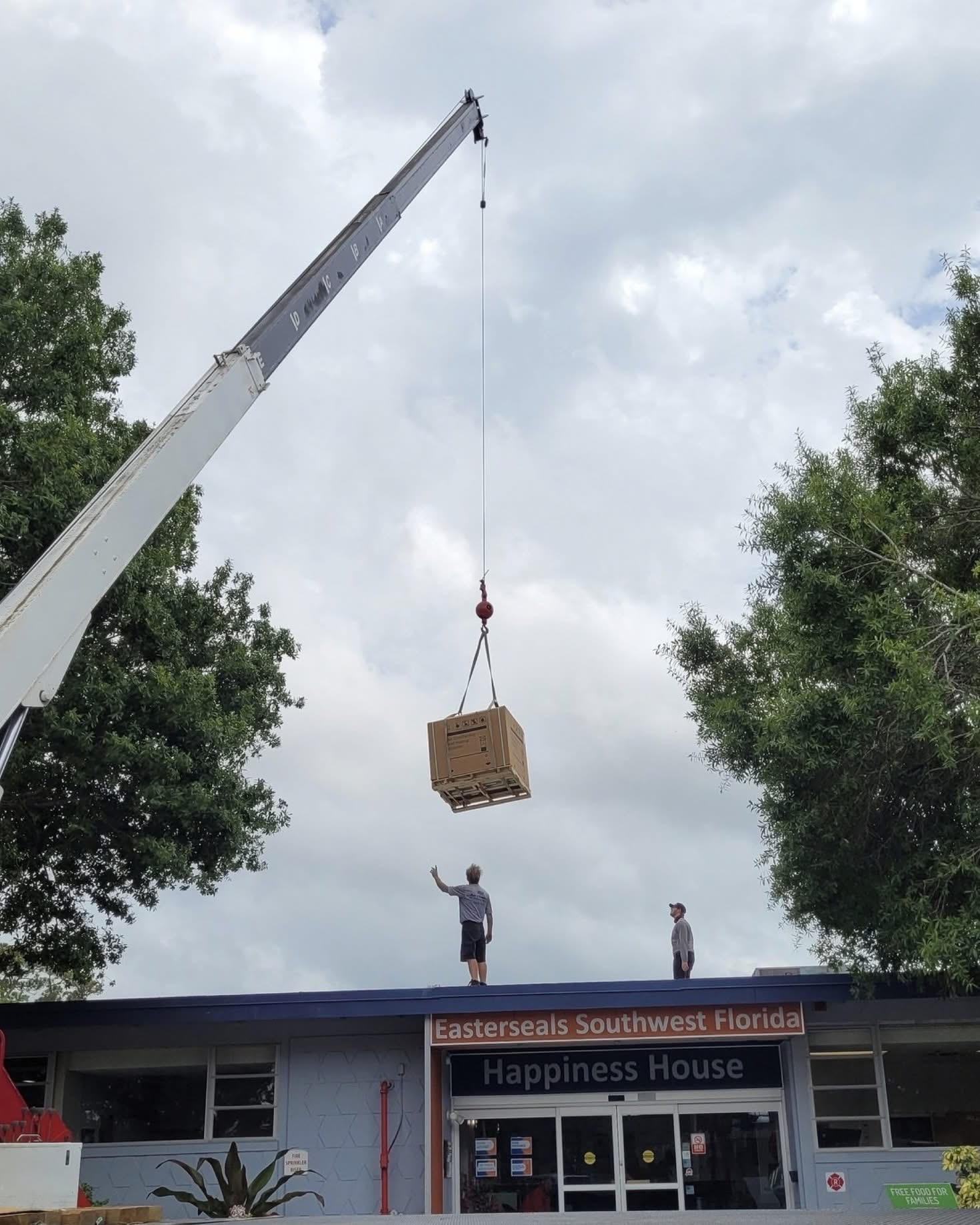 Crane lifting a large cardboard box on a pallet above two people standing on the roof of Easterseals Southwest Florida Happiness House.