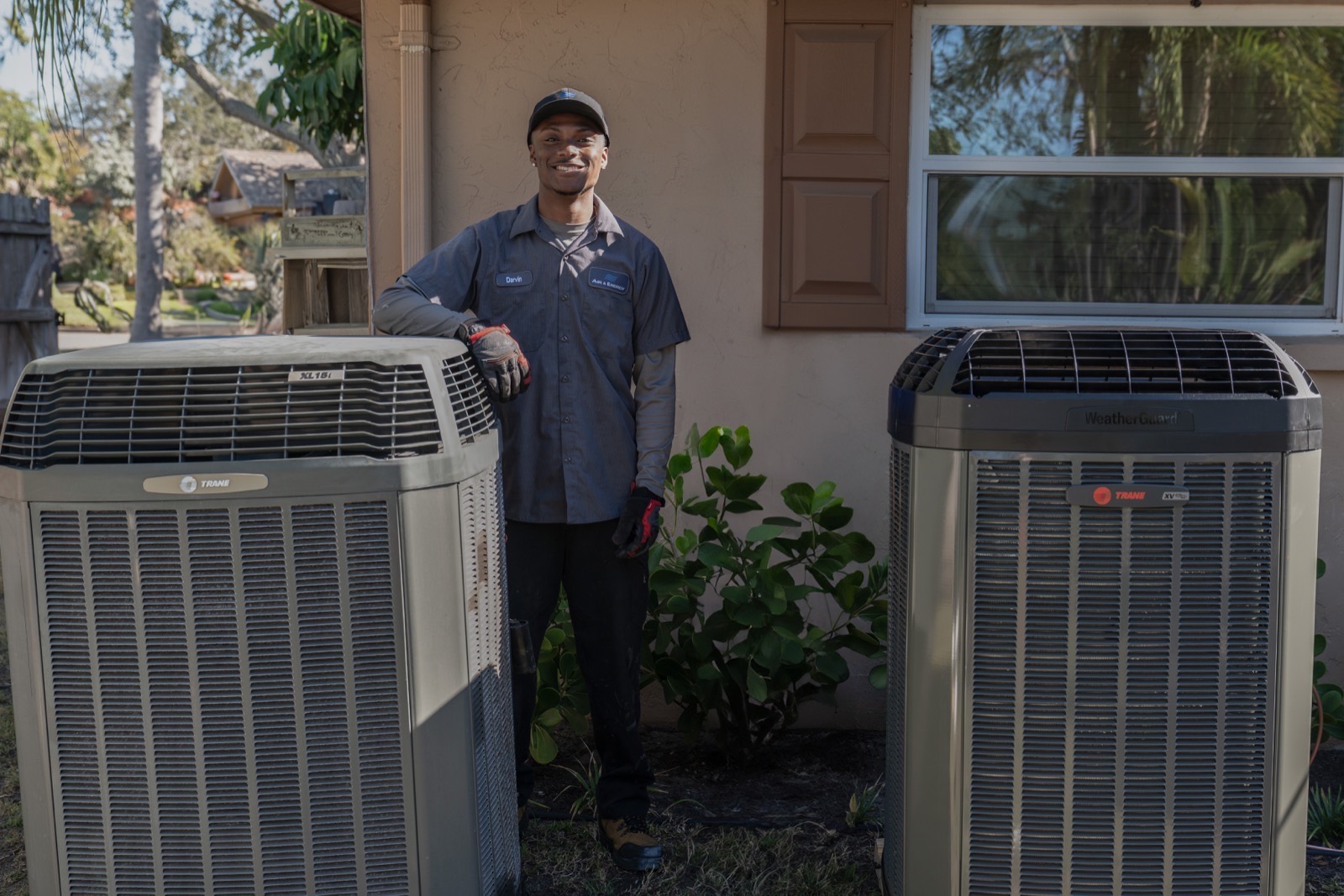An Air & Energy technician standing beside newly installed Trane condensing units in the side yard of a customer's home.