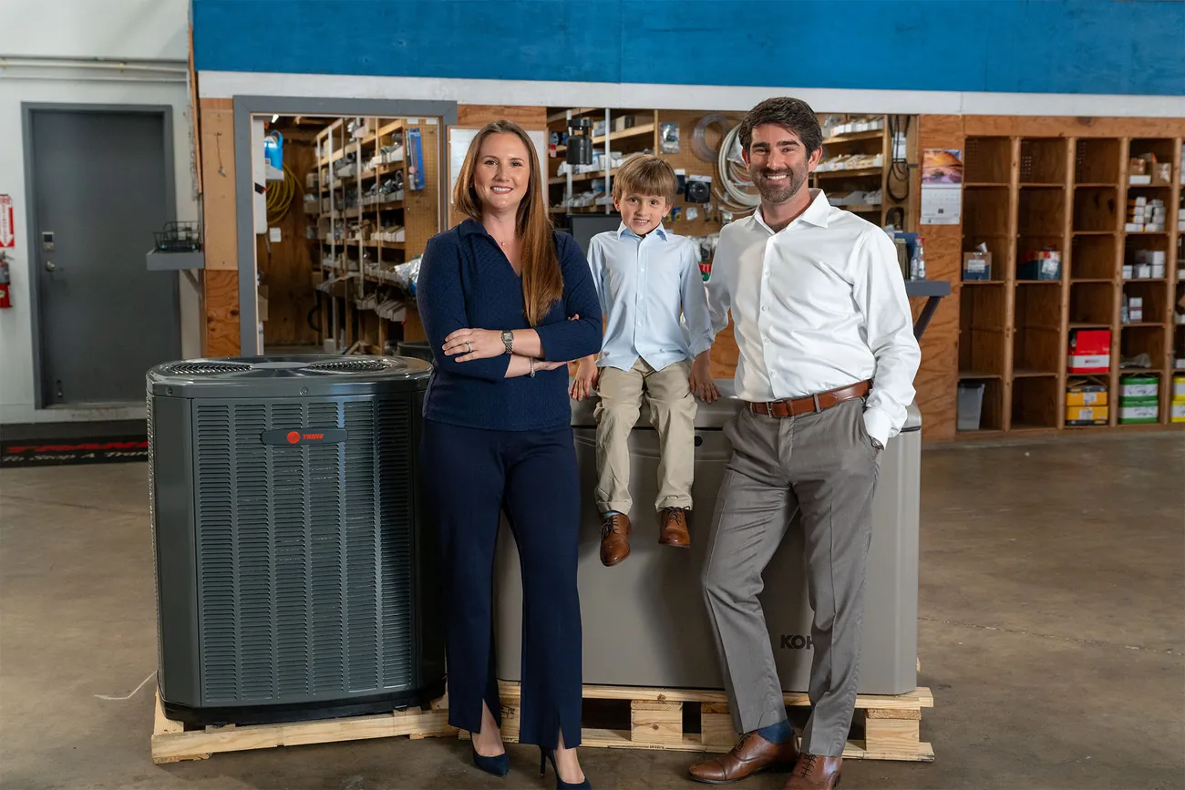 Smiling family of two adults and one child standing in front of a building with a blue stripe and a sign that reads Air & Energy, Air, Plumbing, Electrical.