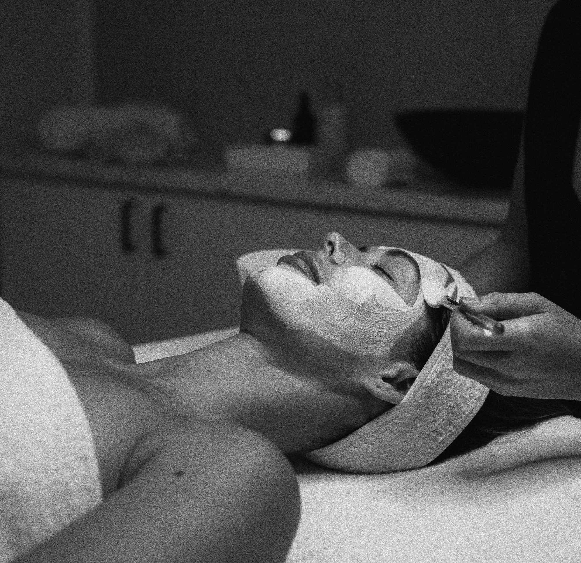 Woman lying down with eyes closed receiving a facial treatment with a brush applying facial mask.