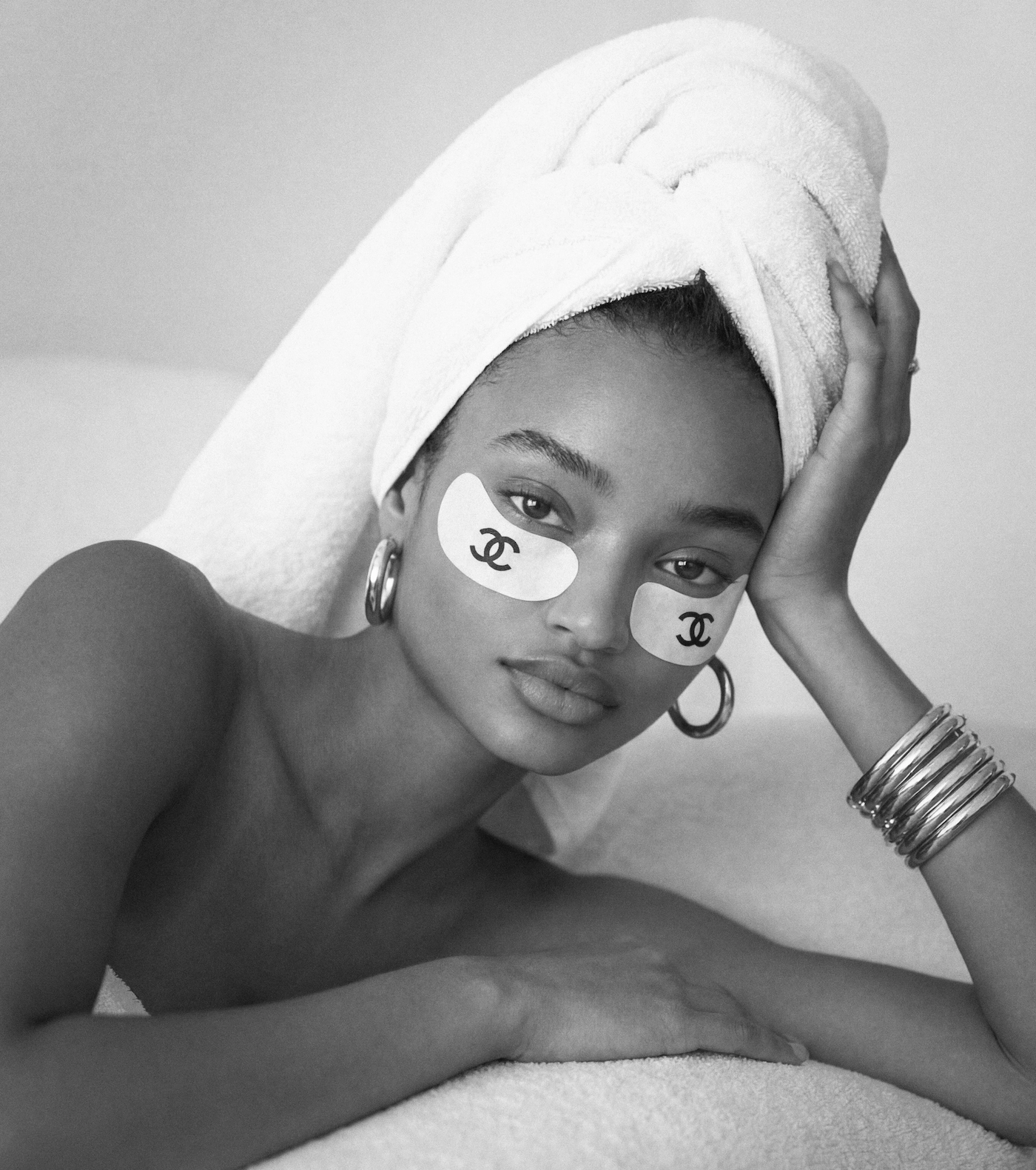 Woman with a towel wrapped around her head wearing Chanel under-eye patches, silver hoop earrings, and bangles.