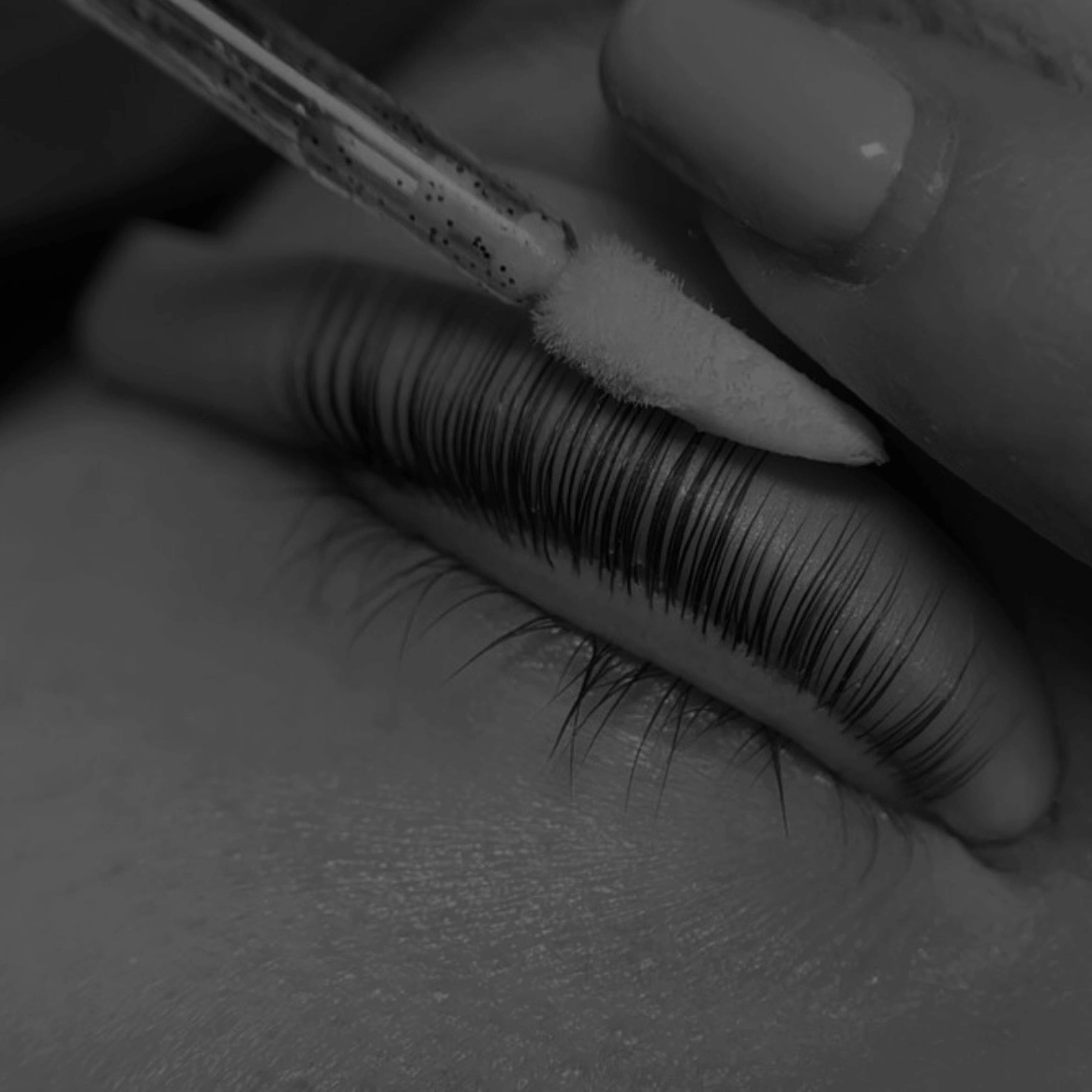 Close-up of a person’s closed eye with long eyelashes and a beauty technician applying a lash lift using a small brush tool.