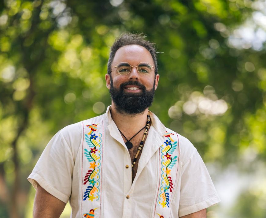 Ryan James Kemp sorri ao ar livre, de óculos e camisa clara bordada, com árvores desfocadas ao fundo.