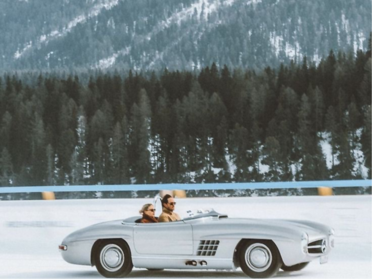 Two people driving a vintage silver convertible car on a snow-covered road with a forest and snowy mountains in the background.