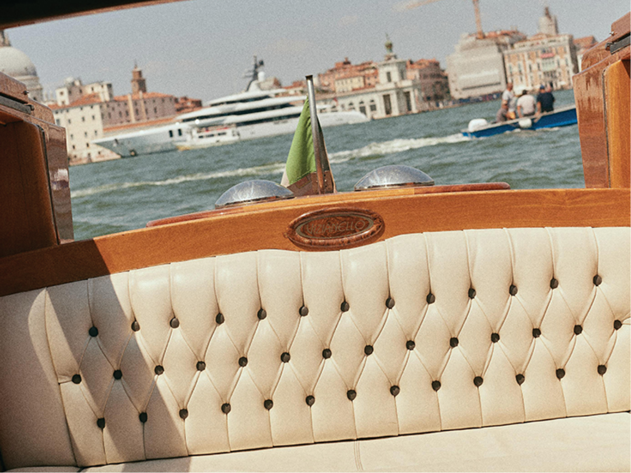 Luxurious beige tufted leather seat on a wooden boat with green flag, water and cityscape in the background.