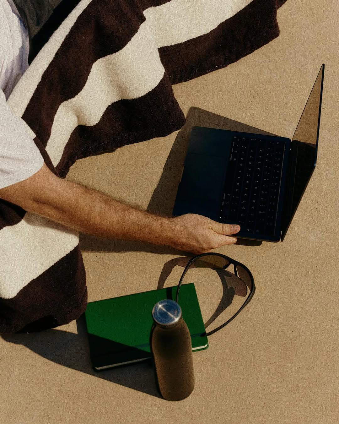 Person holding a laptop while wrapped in a brown and white striped blanket, with a green notebook, sunglasses, and a water bottle on the ground nearby.