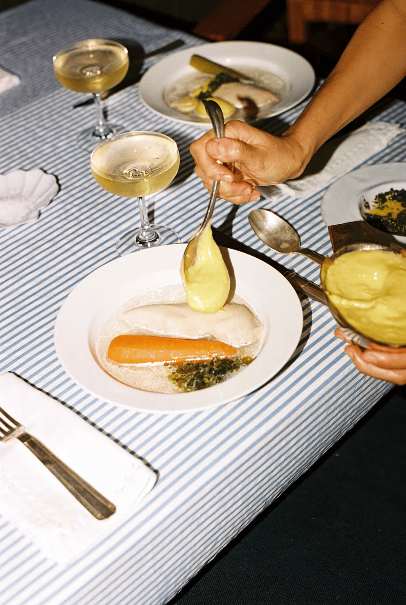 Hand spooning yellow sauce over a plate with boiled chicken, carrot, and herbs on a striped tablecloth alongside two glasses of champagne.
