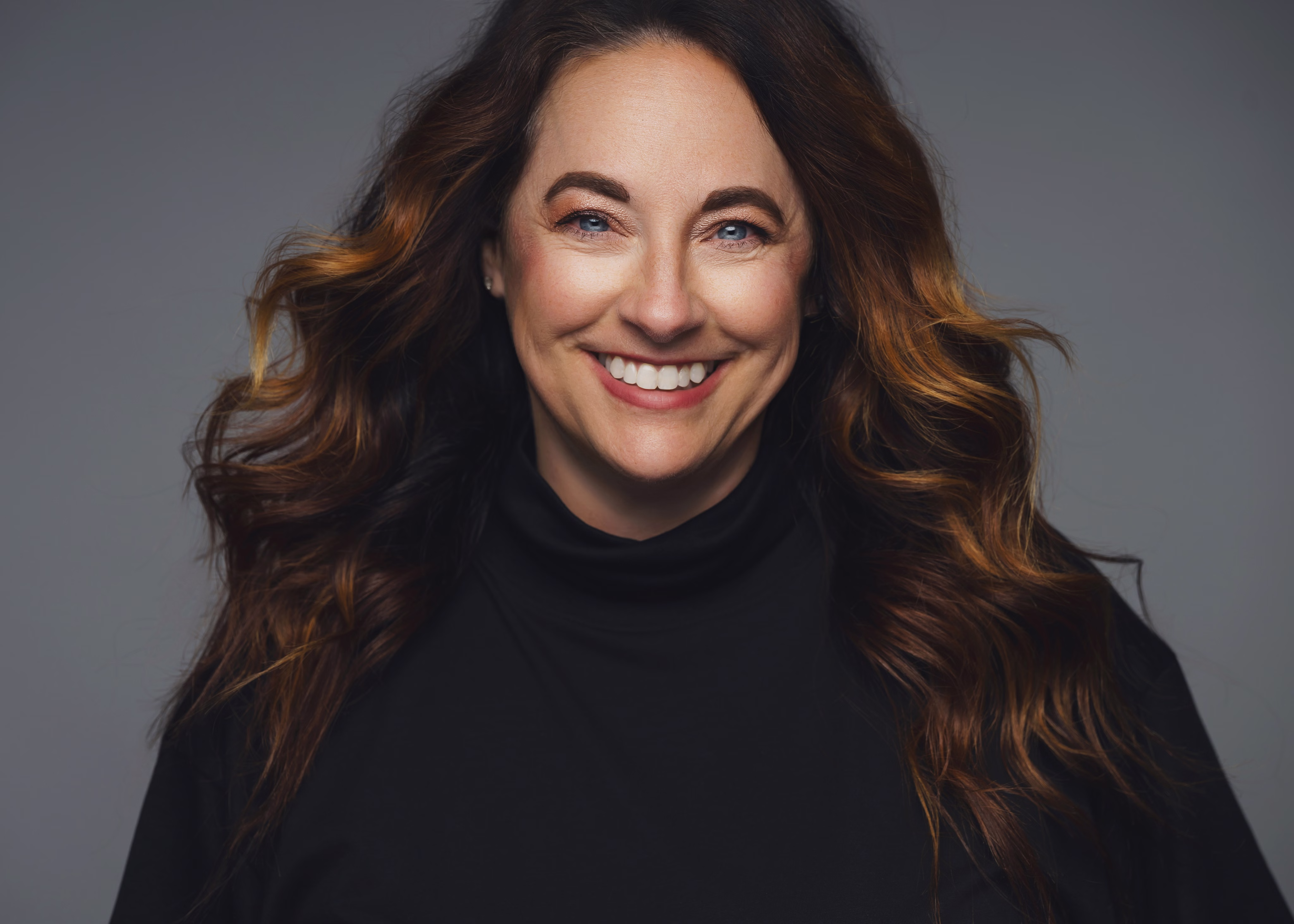 Smiling woman with wavy brown hair wearing a black turtleneck against a gray background.