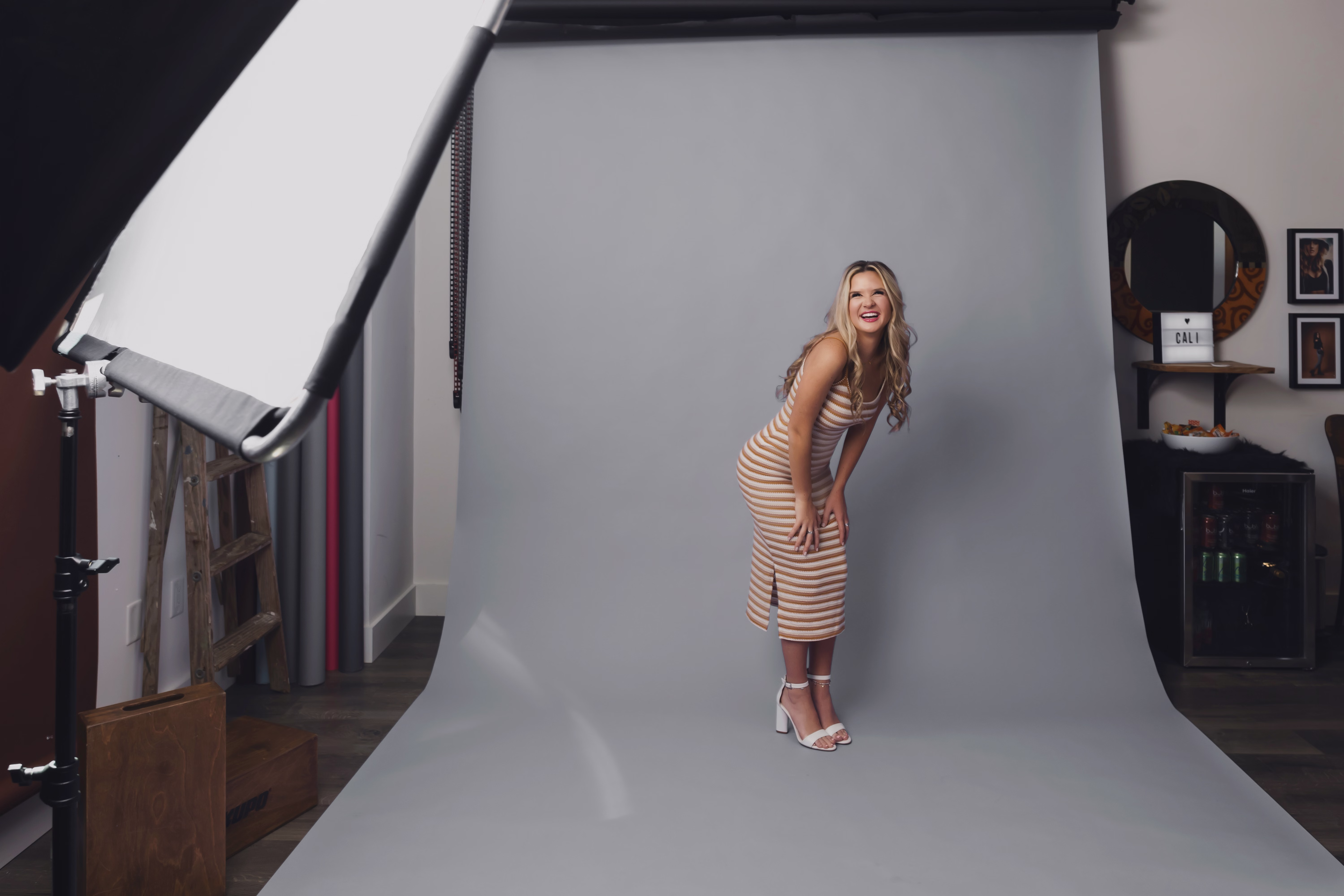 Blonde woman in a striped dress and white heels posing and laughing in front of a gray photography backdrop in a studio.