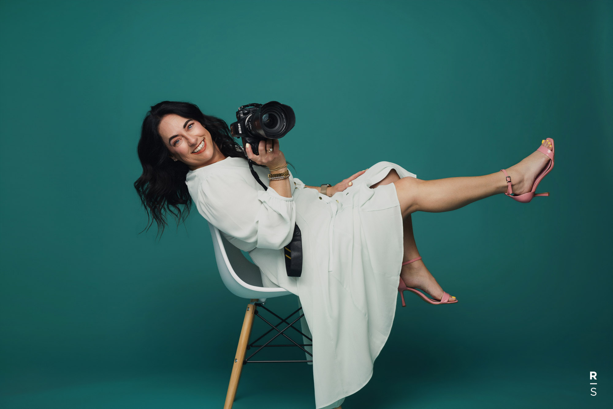 Smiling woman in white dress sitting sideways on chair holding a Nikon camera with legs extended.