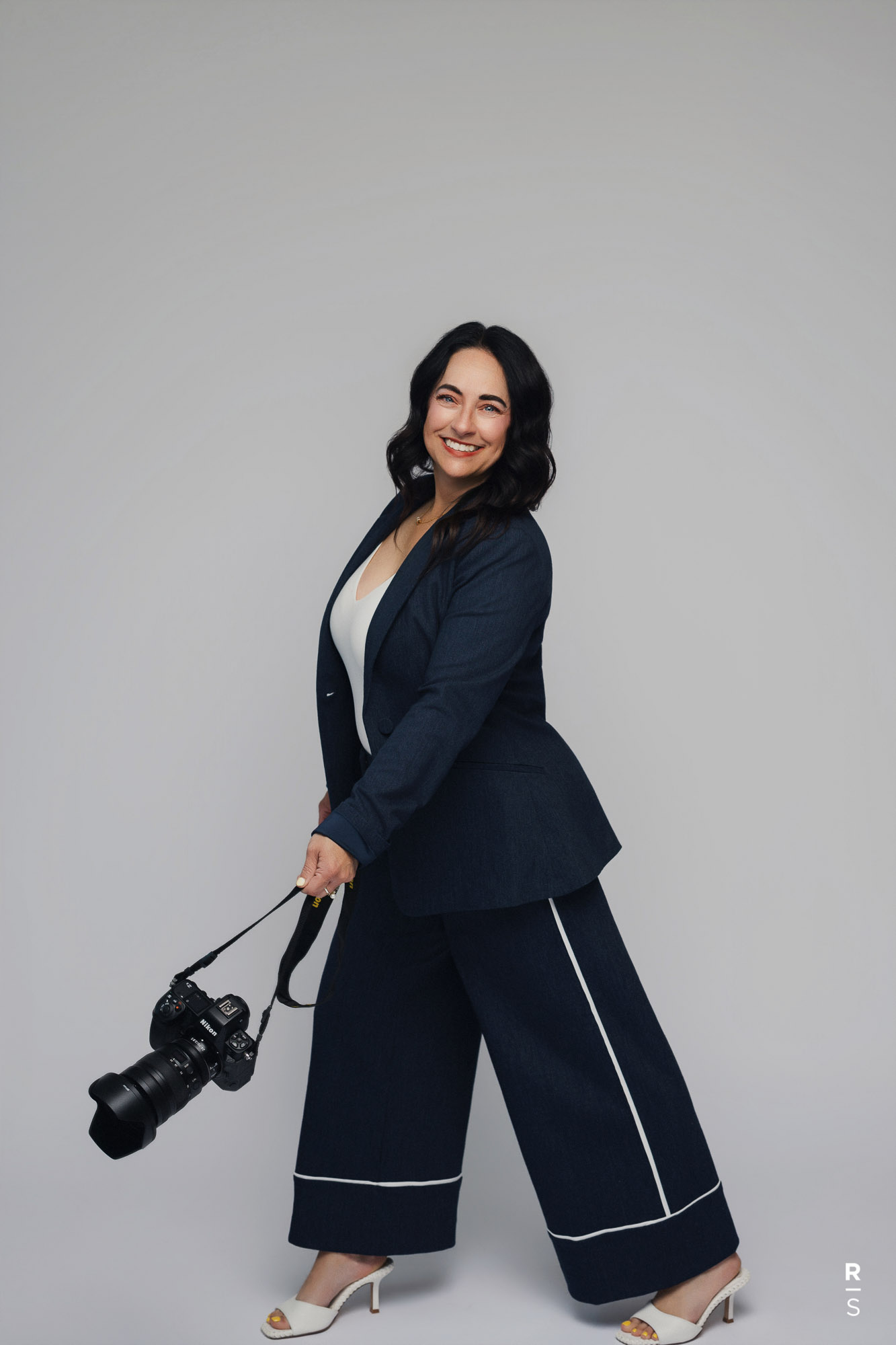 Smiling woman in navy suit holding a camera walking against a plain gray background.