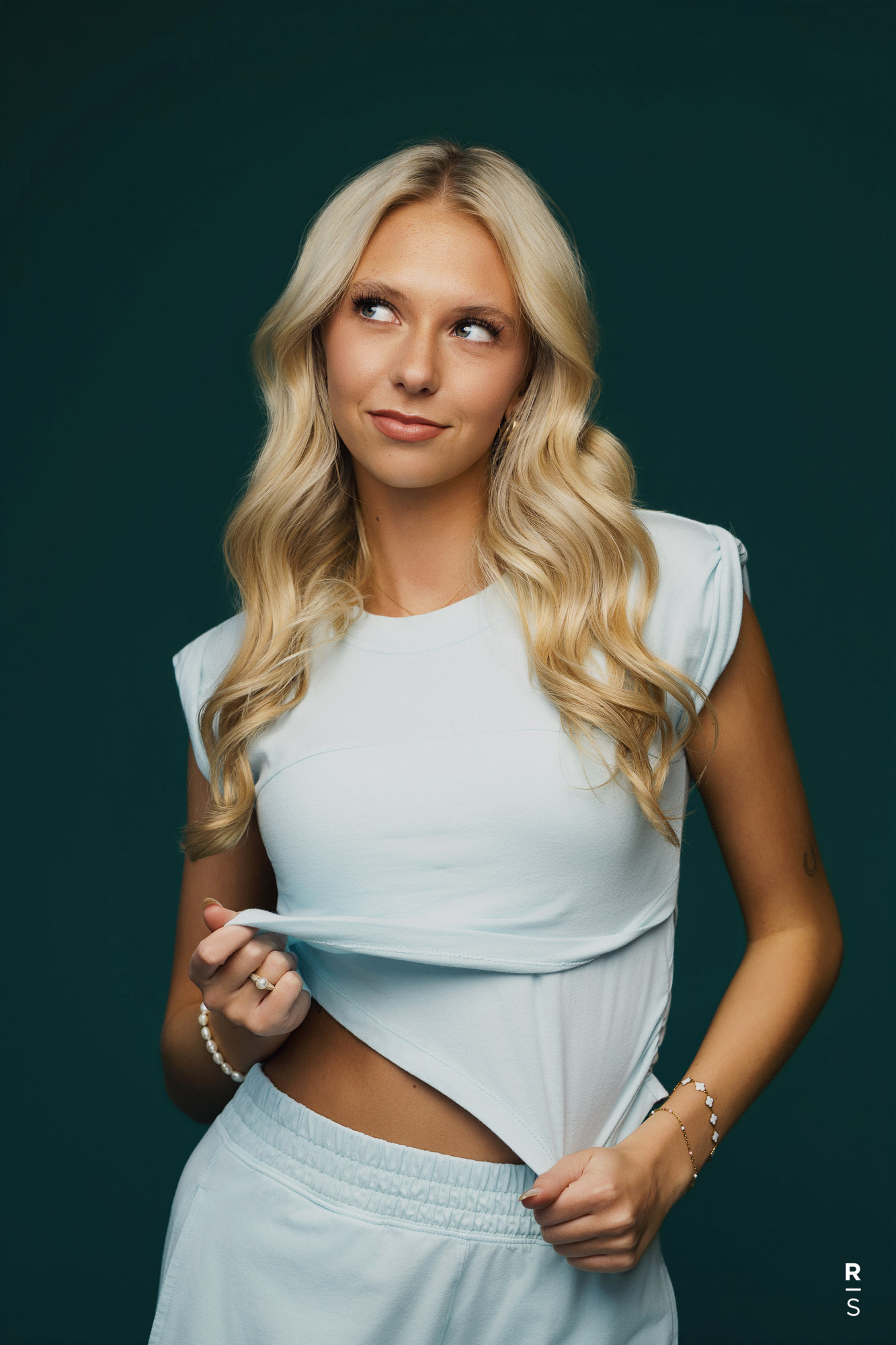 Blonde woman in light blue outfit lifting shirt slightly and looking up to her left.