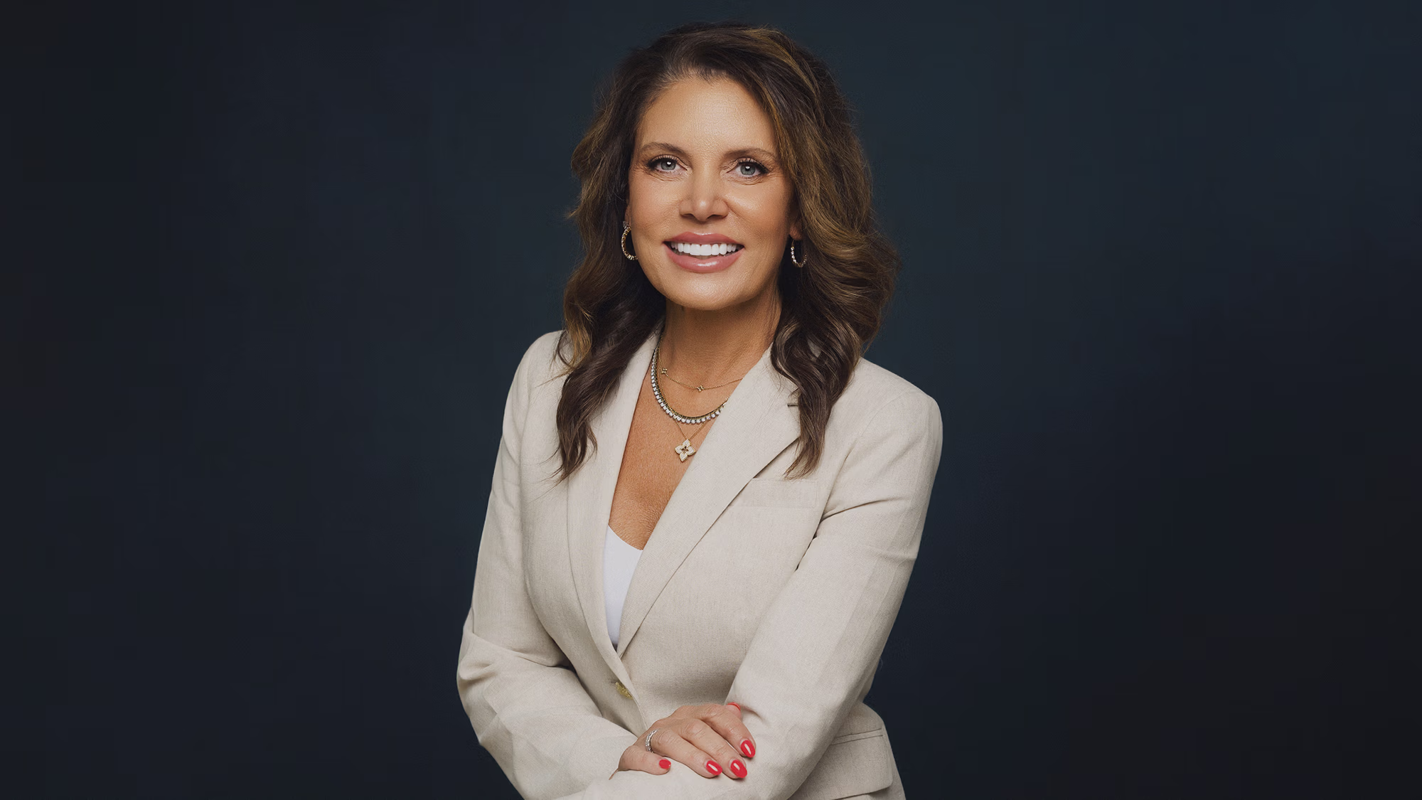 Smiling woman with wavy brown hair in a beige blazer and red nail polish against a dark background.