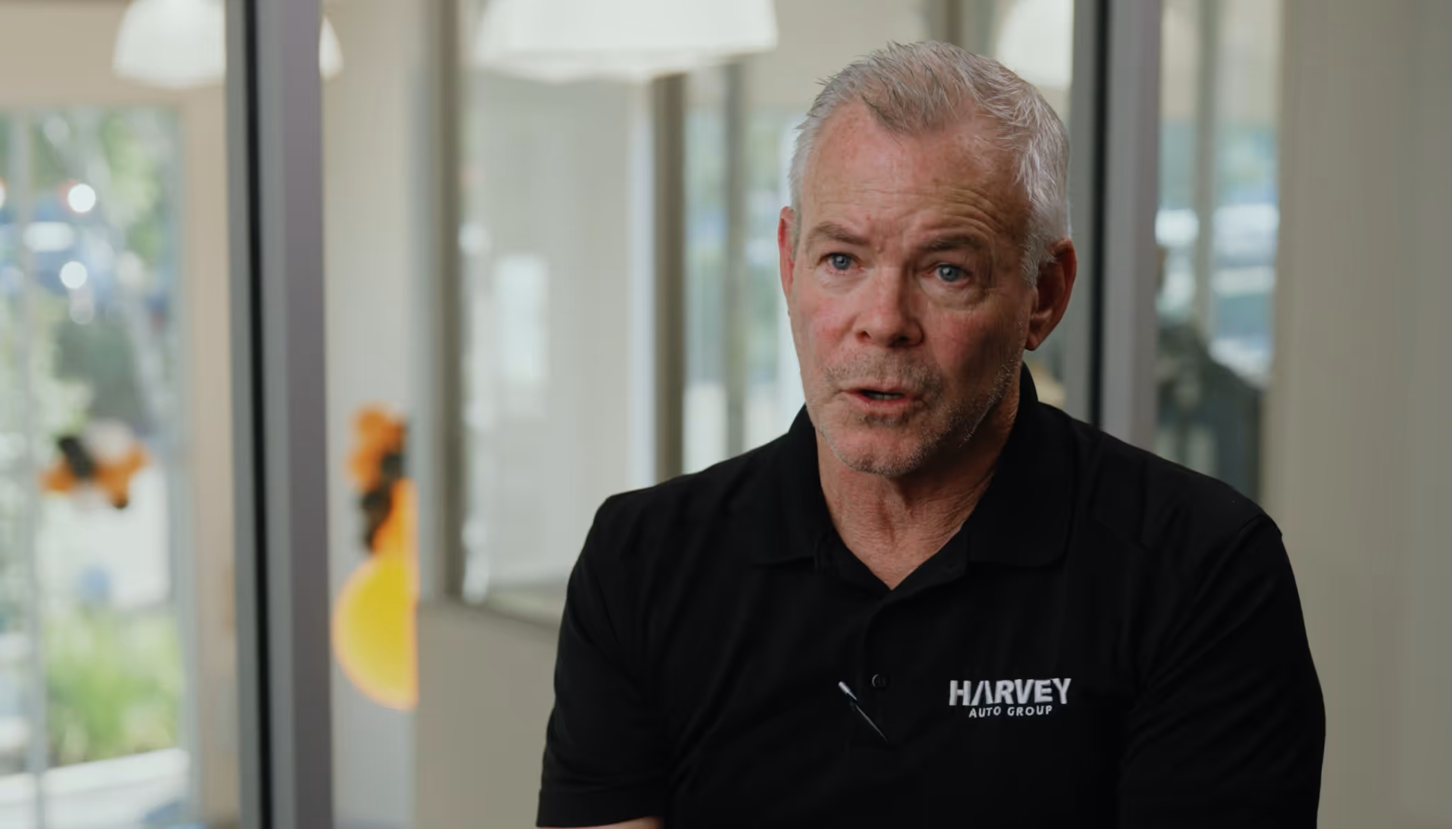 Middle-aged man wearing a black Harvey Auto Group polo shirt speaking in a bright office setting.