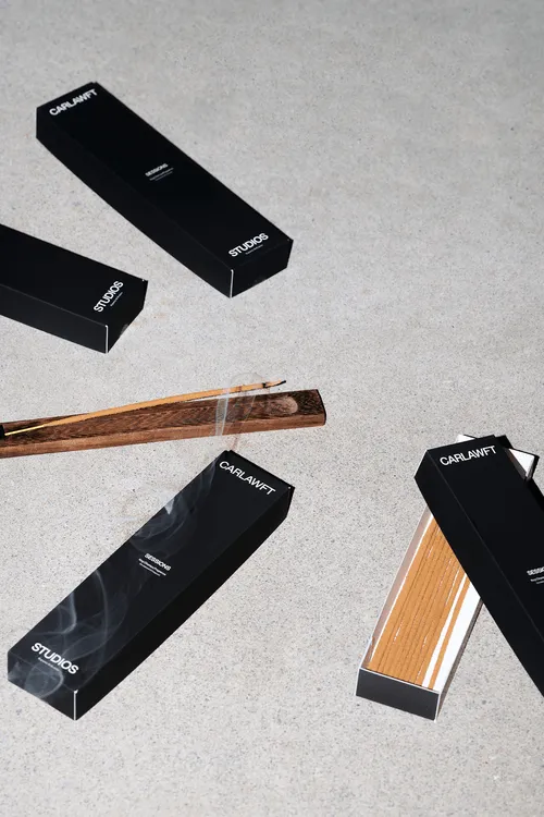 Black CARLAWFT Studios incense boxes on a grey surface with one incense holder holding a burning incense stick emitting smoke.