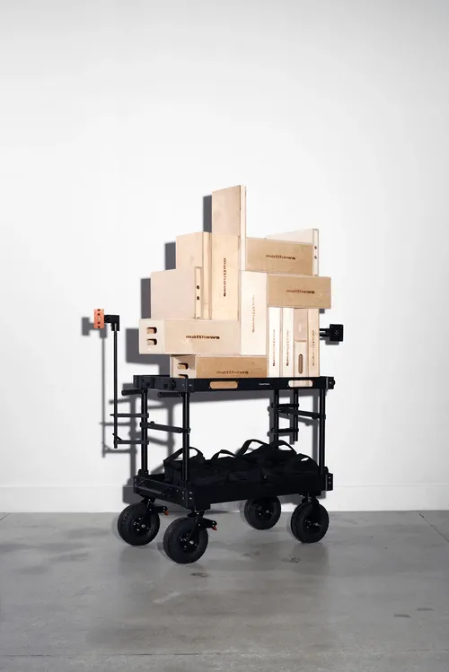 Black utility cart with large wheels holding stacked wooden Matthews brand boxes against a plain white wall.