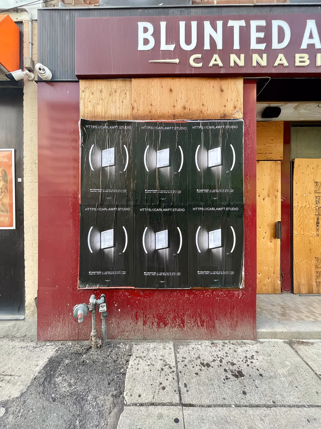 A maroon storefront with boarded-up windows covered by six identical black posters advertising carlawft.studio, showing a blurred white image inside parentheses.