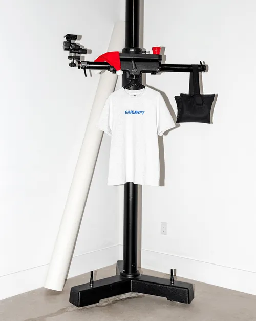 White t-shirt with blue text 'CARLAWFT' hanging from a black tripod stand with a red Santa hat and small red cup on top.