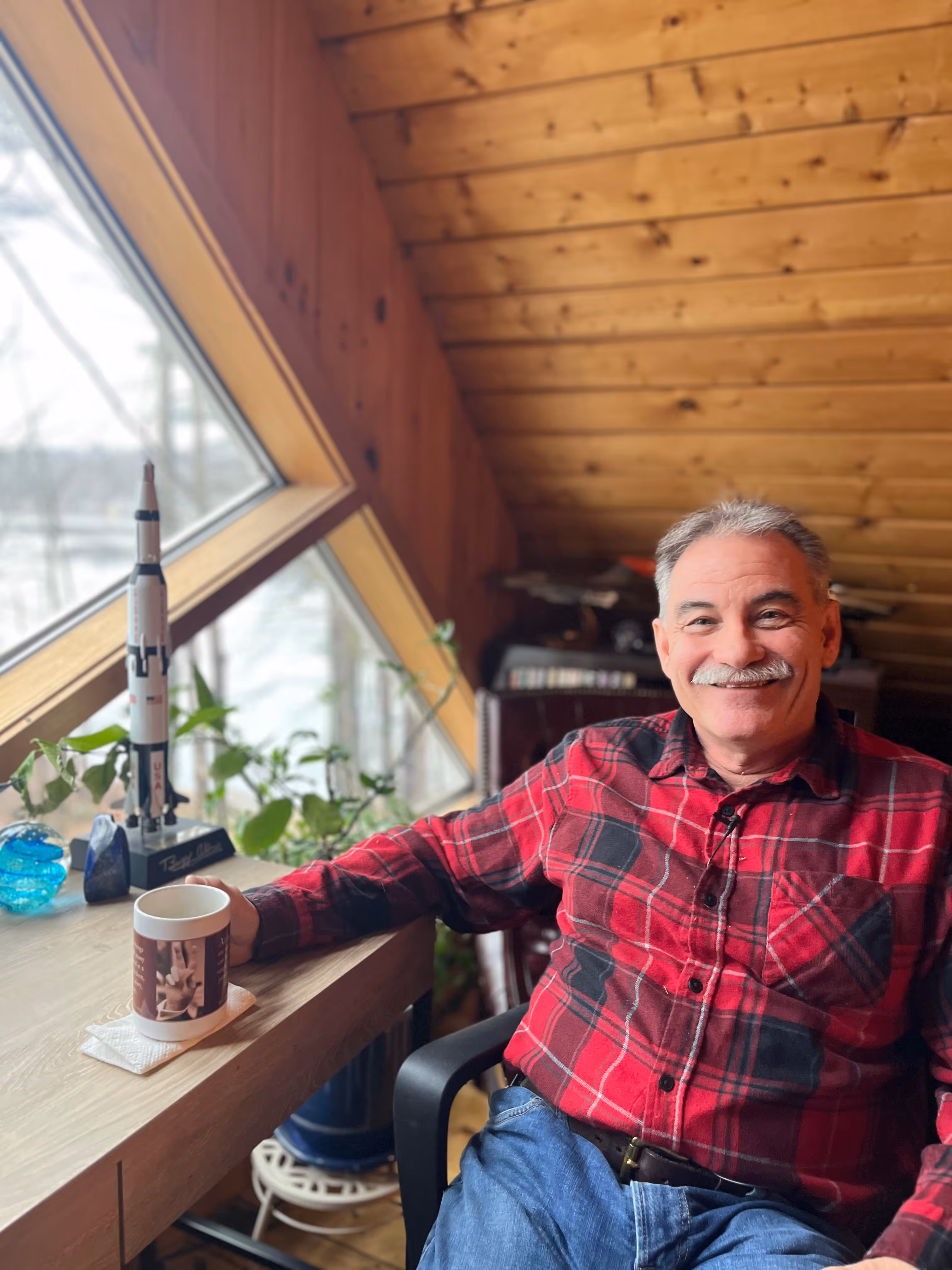Smiling man in a red plaid shirt sitting by a wooden table.